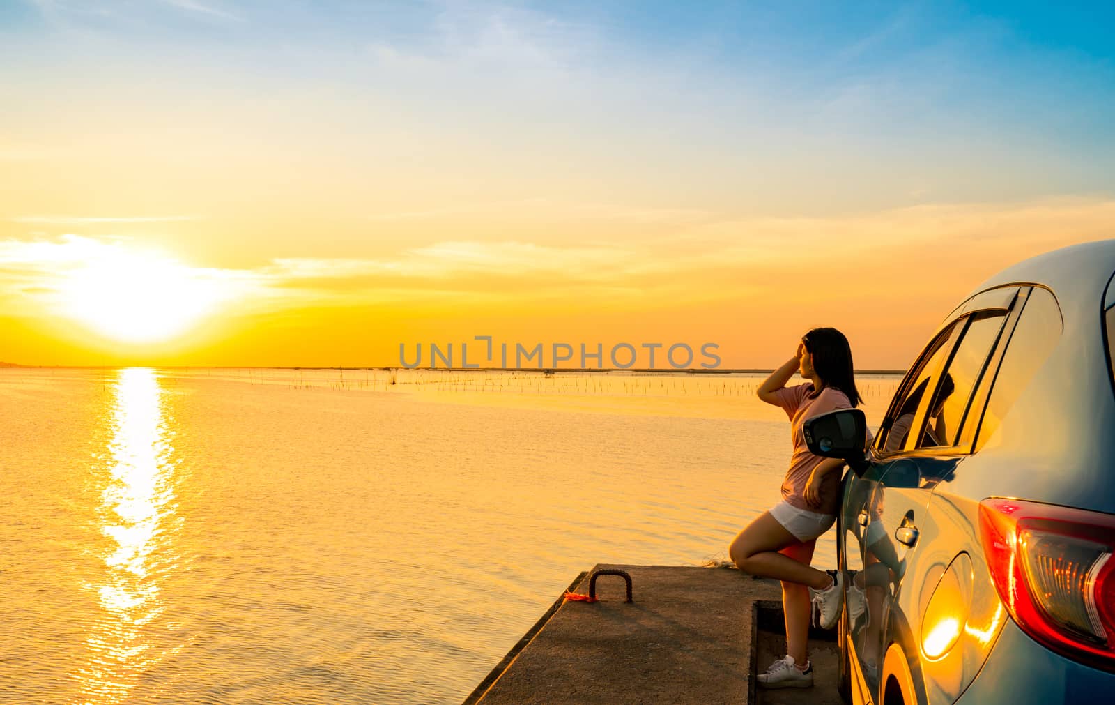 Young Asian woman wear T-shirt, short pants stand leaning blue compact SUV sport car by the sea at sunset with beautiful clouds and yellow-orange sky and reflection on water