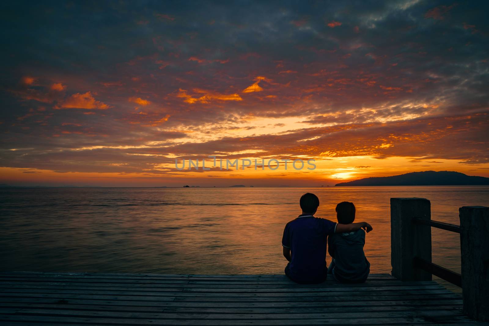 Silhouette of young romantic couple in love is sitting and hugging on wooden pier at the beach in sunrise time with golden sky. Vacation and travel concept. Romantic young couple dating at seaside. by Fahroni