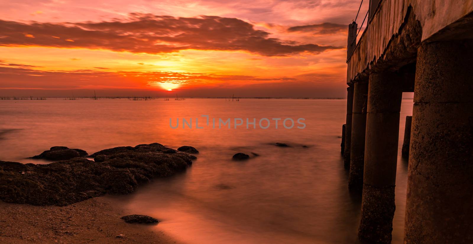 A long exposure landscape of beautiful sunset at the sea beach near concrete bridge with orange sky, clouds and stone by Fahroni