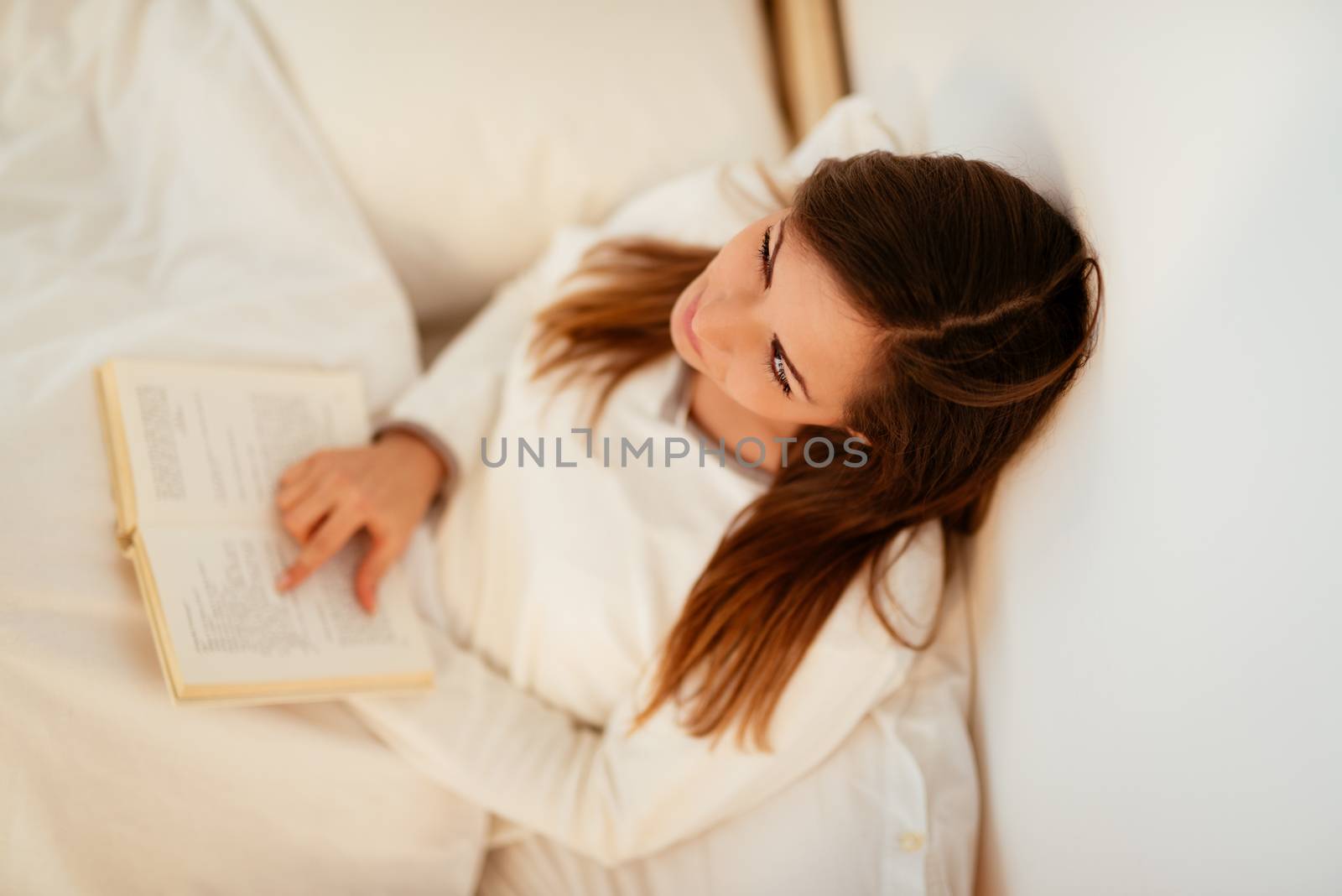 Beautiful smiling pensive girl relaxing in bed and reading book. Top view.