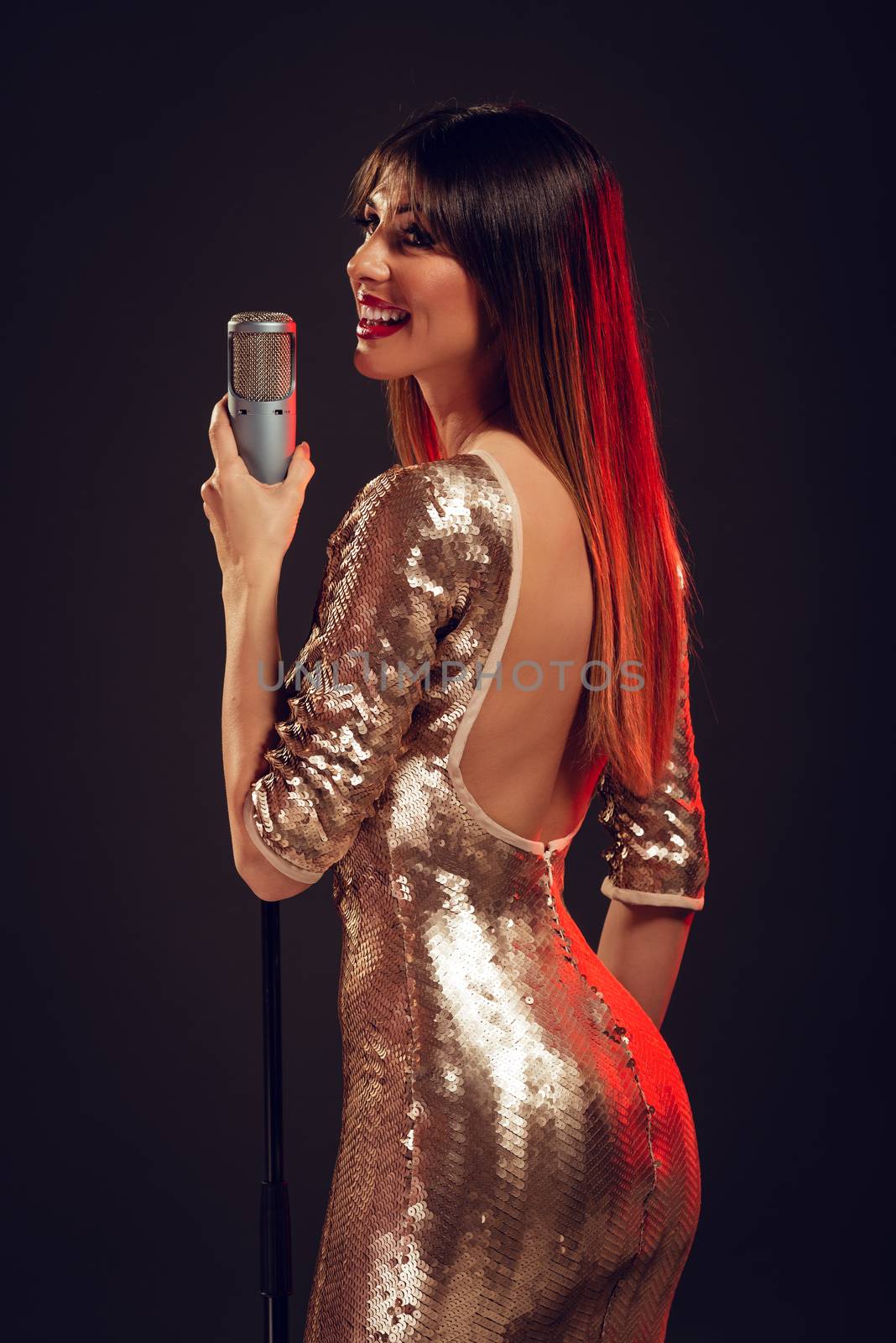 A young smiling woman in sequin dress singing in front of a microphone with expression of happiness on her face.