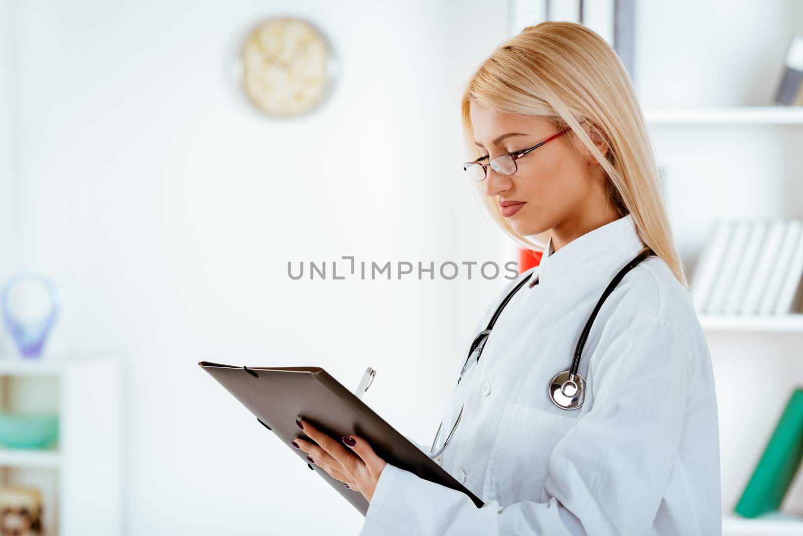 Young cute female doctor with the glasses, dressed in a white coat standing and holding folder with a stethoscope over the neck.