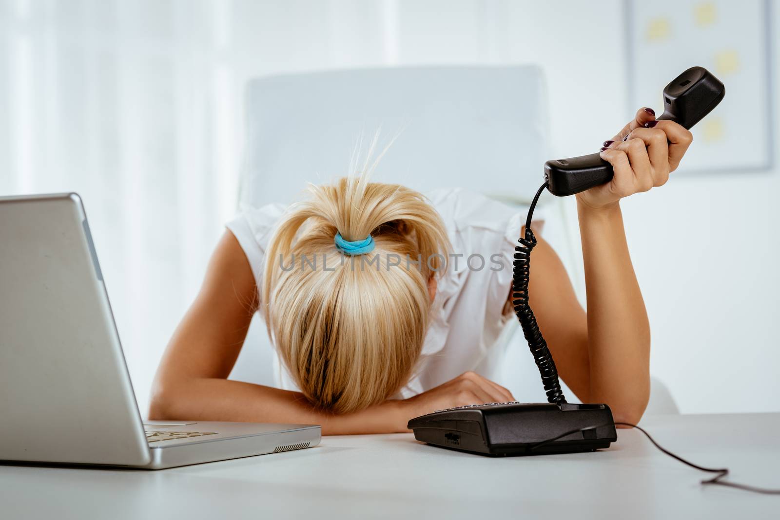 Young business woman napping in the office, on her desk, with a telephone handset on hand, next to laptop.