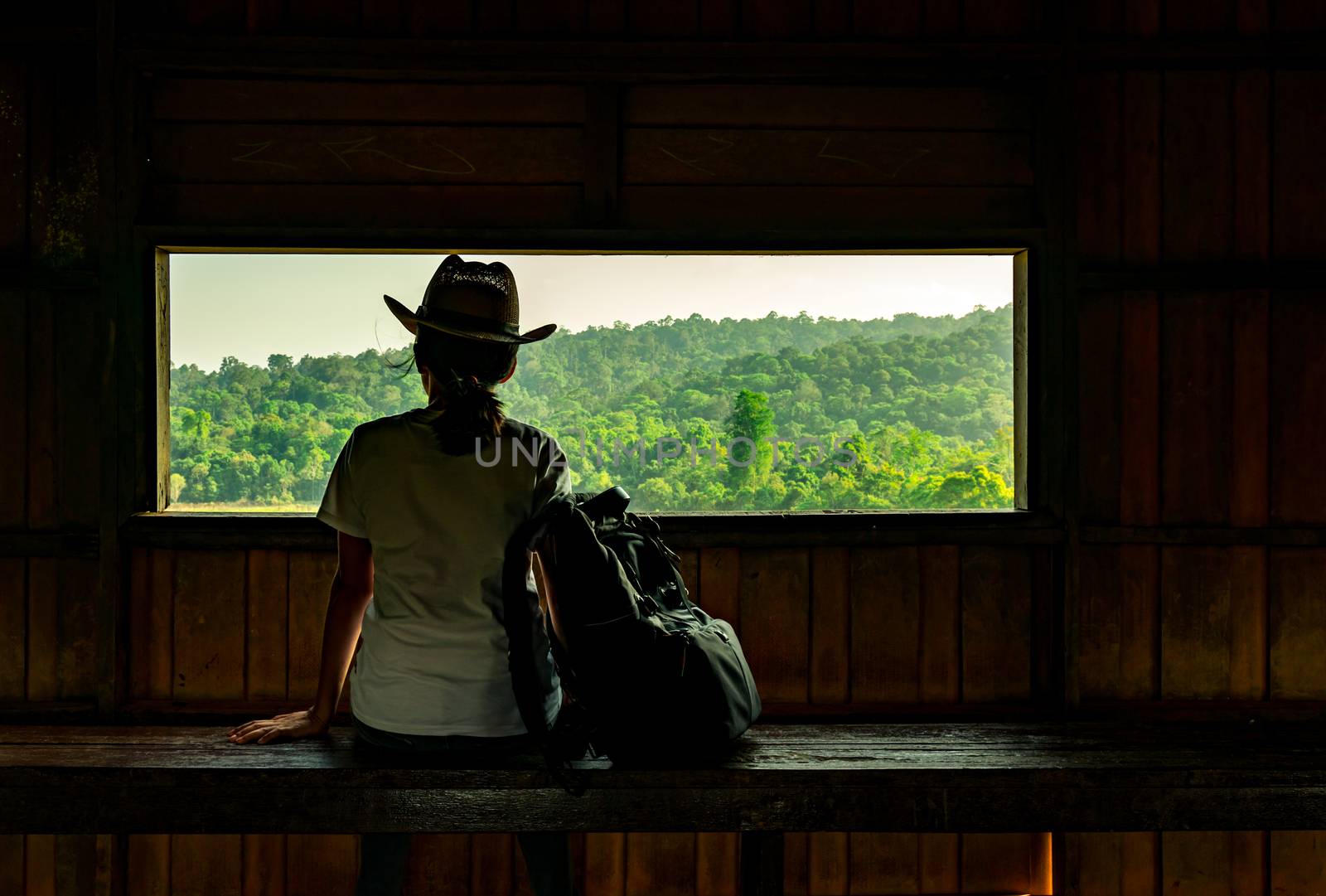 Young Asian woman wear the hat sit on wooden bench and watching beautiful view of tropical forest at wildlife observation tower in the evening with warming sunlight. Woman travel alone concept.