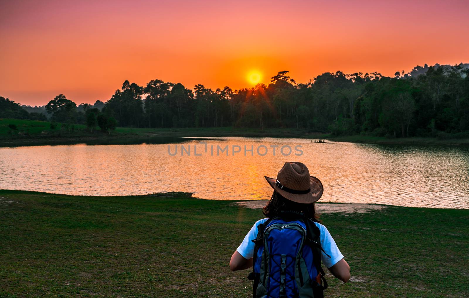 Woman tourist with hat and backpack standing with relax mood and watching sunset behind the pond and evergreen forest. Alone young woman traveller enjoy beautiful scenery sunset. by Fahroni