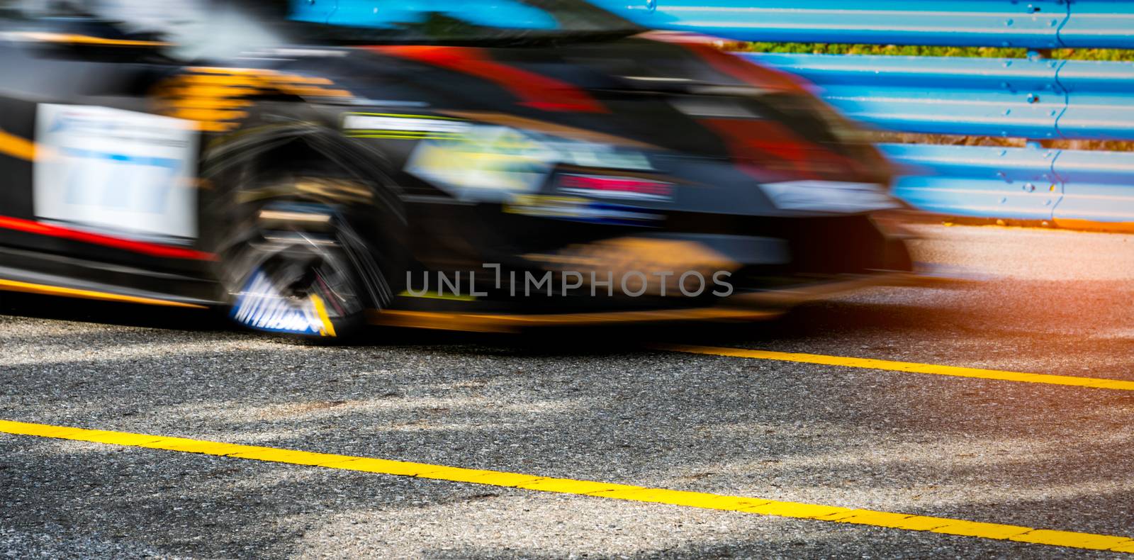 Motor sport car racing on asphalt road with blue fence and yellow line traffic sign. Car with fast speed driving and motion blurred. Black racing car with red and yellow stripes. Car on racetrack.