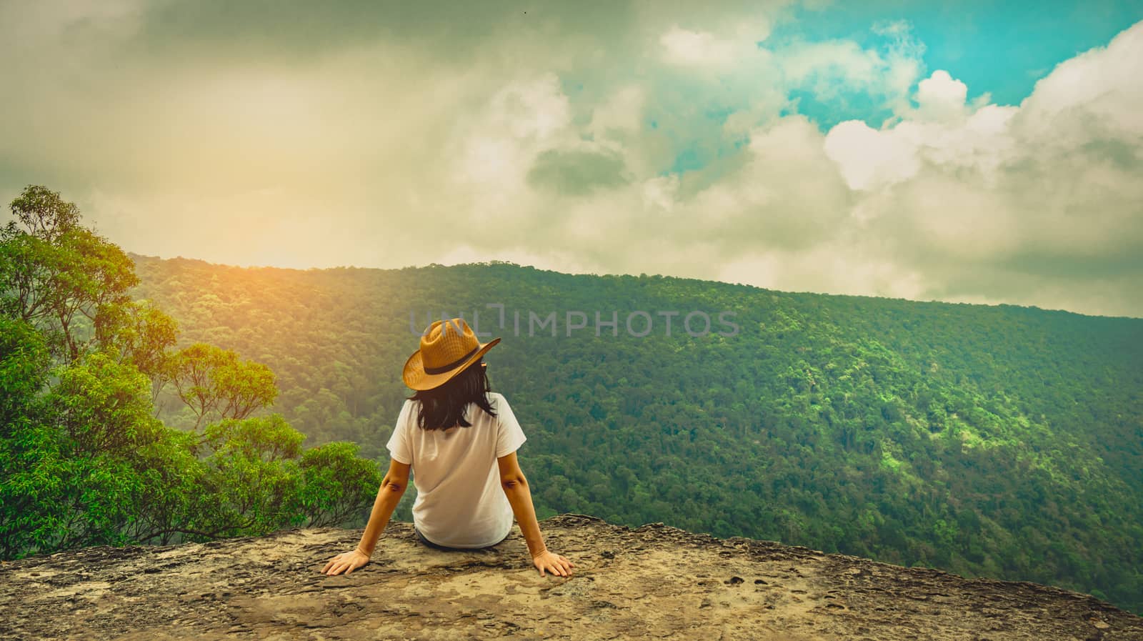 Young traveling woman wearing hat and sitting on the top of the mountain cliff with relaxing mood and watching beautiful view of woods and blue sky and clouds on vacation. Asian woman travel alone. by Fahroni