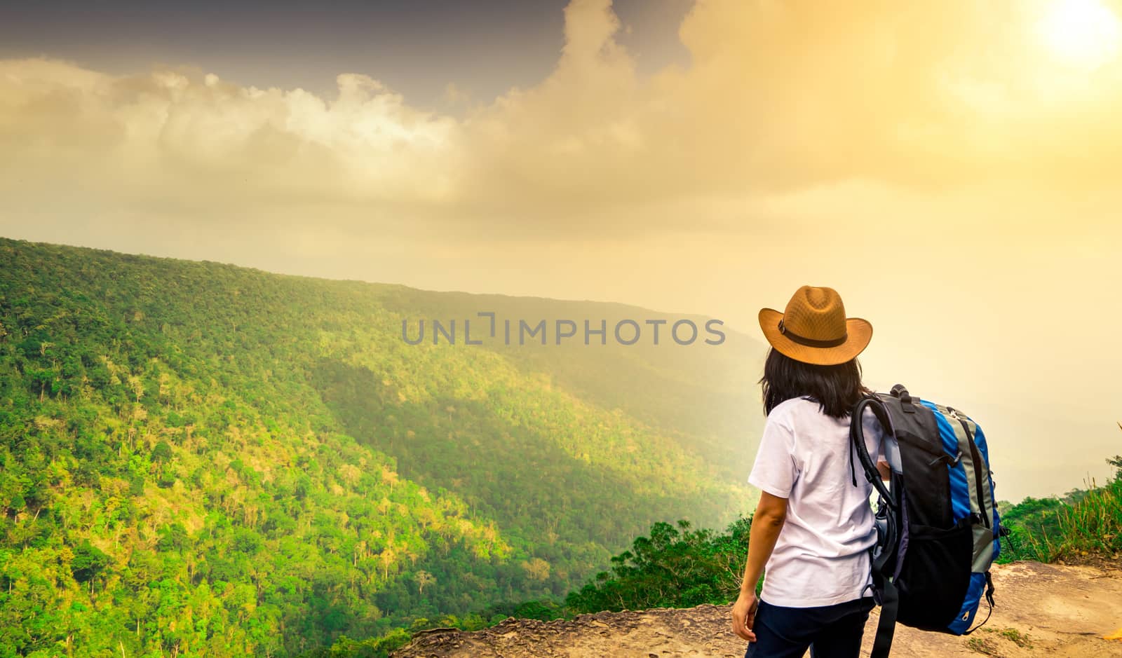 Young traveling woman with backpack and hat stand on the top of the mountain cliff watching beautiful view of woods and sky after rain on her vacation. Asian woman travel alone. by Fahroni