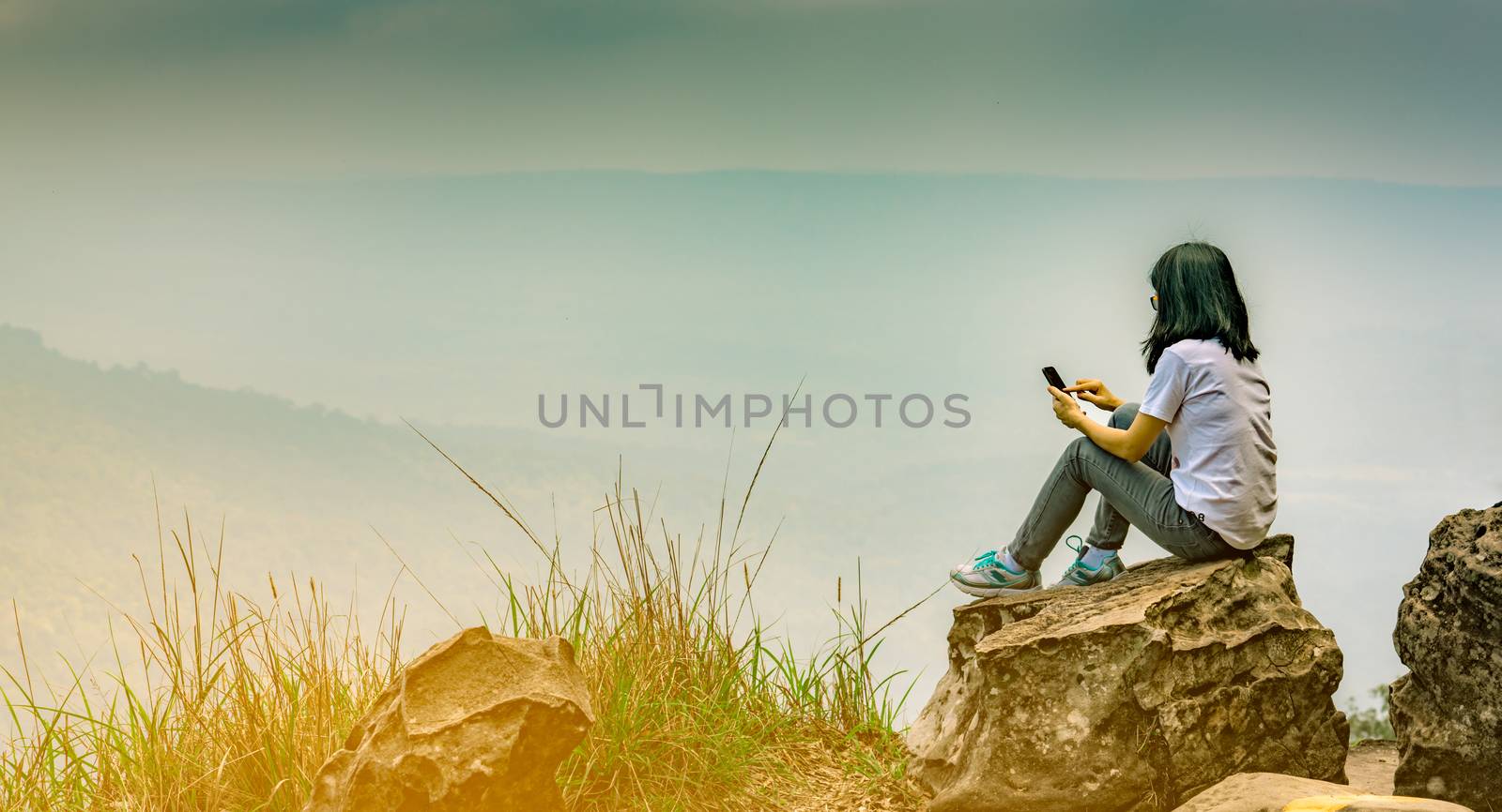 Young Asian woman sit on the rock at the cliff and using smart phone message to her friends while on vacation at the national park. Adventure travel and communication concept