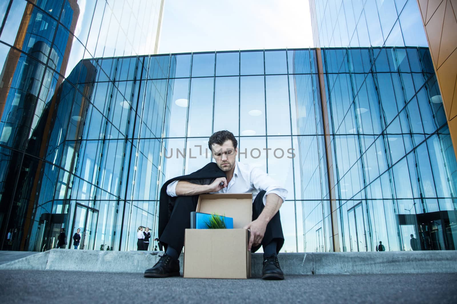 Fired business man sitting frustrated and upset on the street near office building with box of his belongings. He lost work