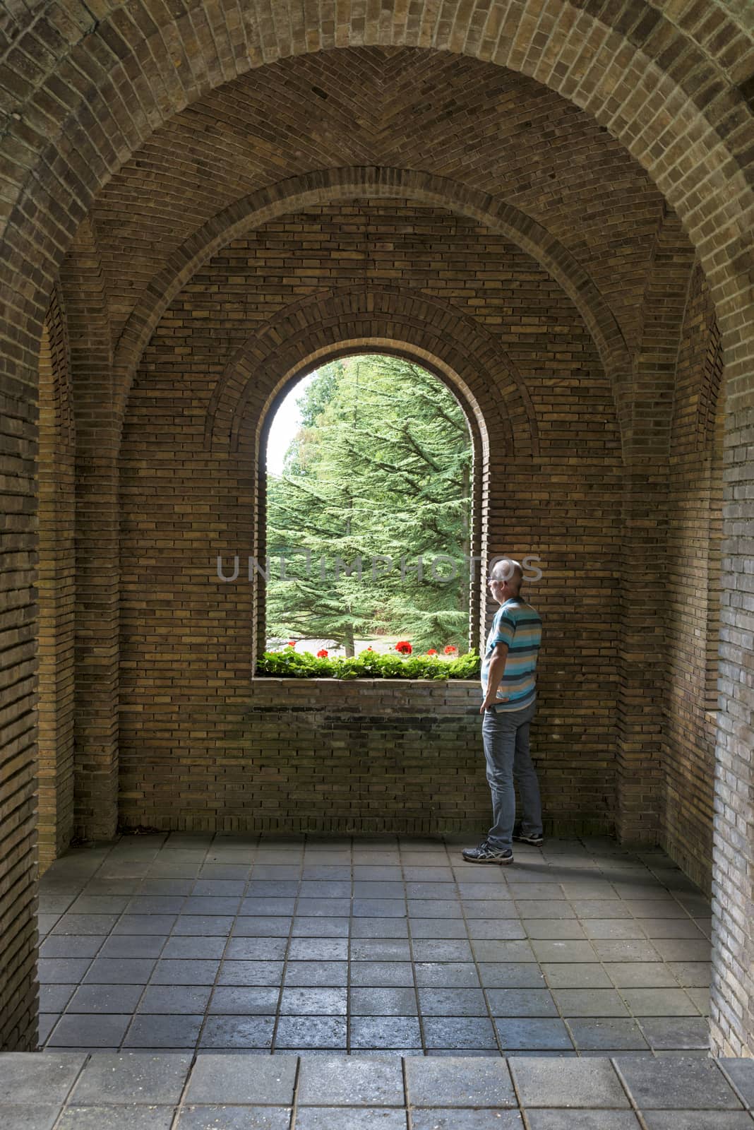 Nijmegen,Holland,26-August-2018:adult man looking at the nature from a old stone corridor with see trhough in old building with green plants and red flowers in the wondow opening