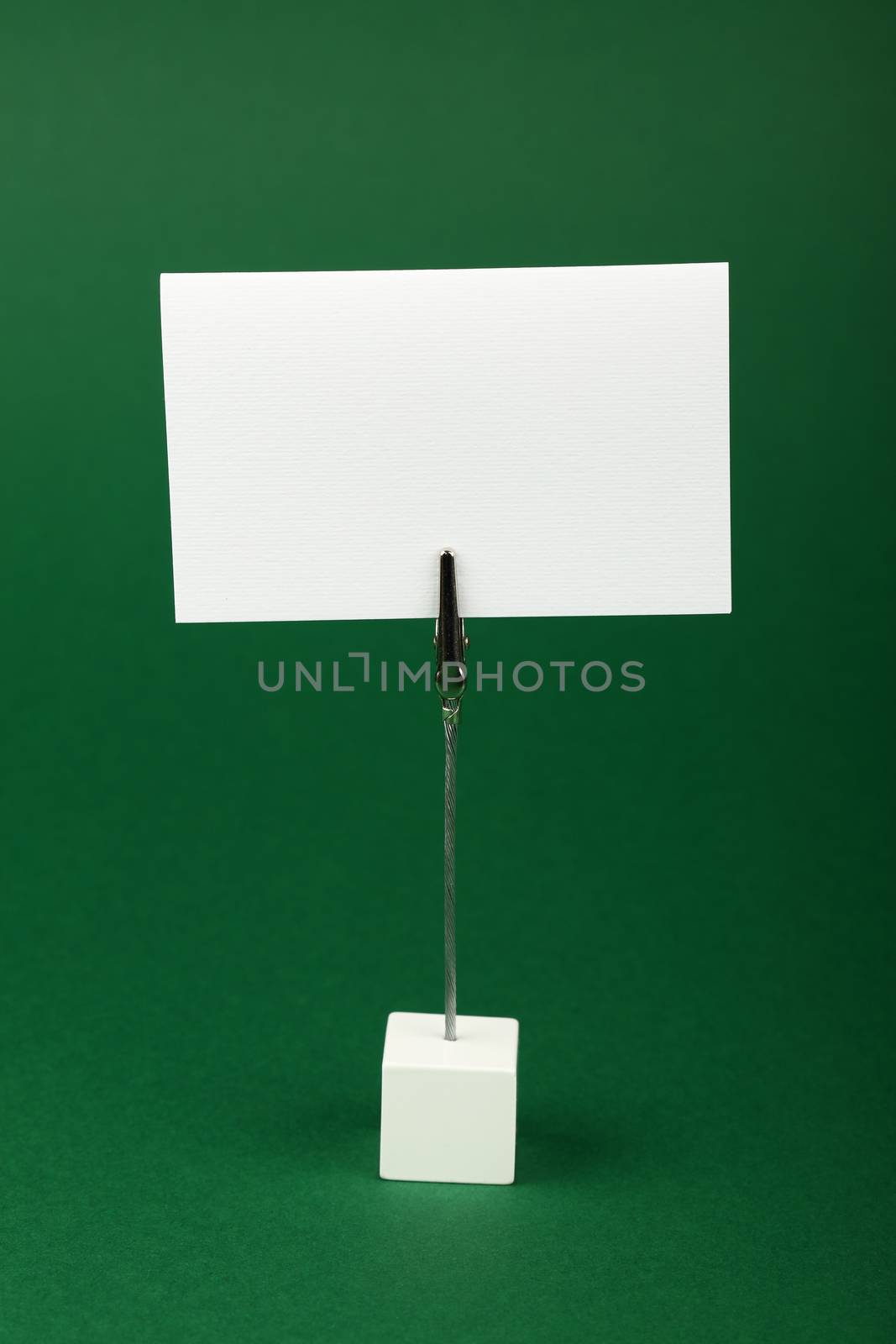 Blank white cardboard sign with copy space on metal note holder over green, paper background, front side view