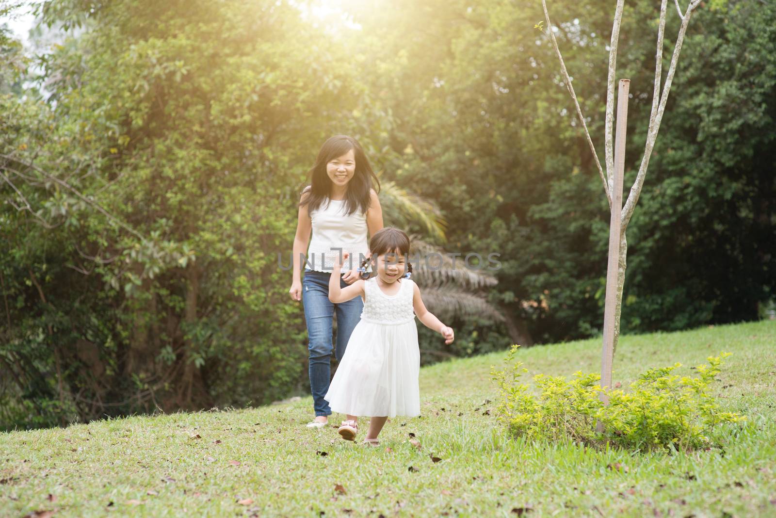 Mother and little girl play chasing at green park. Asian family outdoors activity.