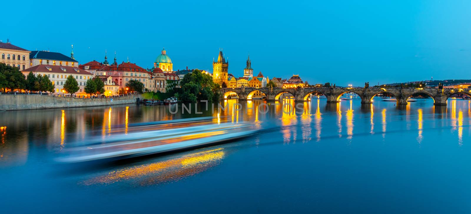 Illuminated Charles Bridge reflected and blurred tourist boat in Vltava River. Evening in Prague, Czech Republic.