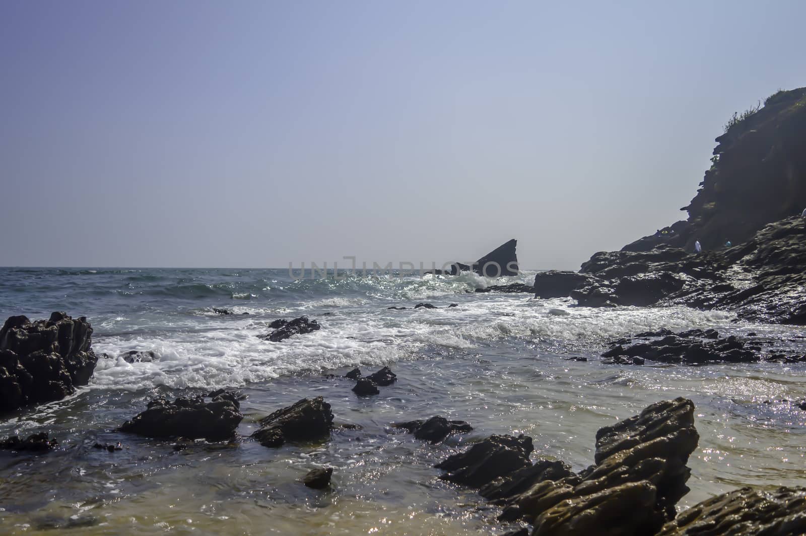Sea beach on bay of Indian Ocean, Goa, India. Emerald waves of ocean under blue sky. Rocky coastline ends at sea shore and mountain. The coastline is filled with rocks and blue waves at sunset.