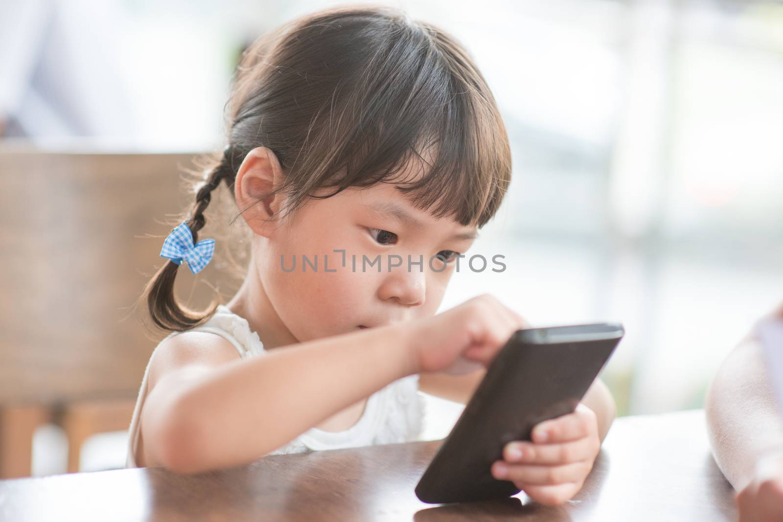 Little Asian girl using smart phone at cafe. Natural light outdoor lifestyle.