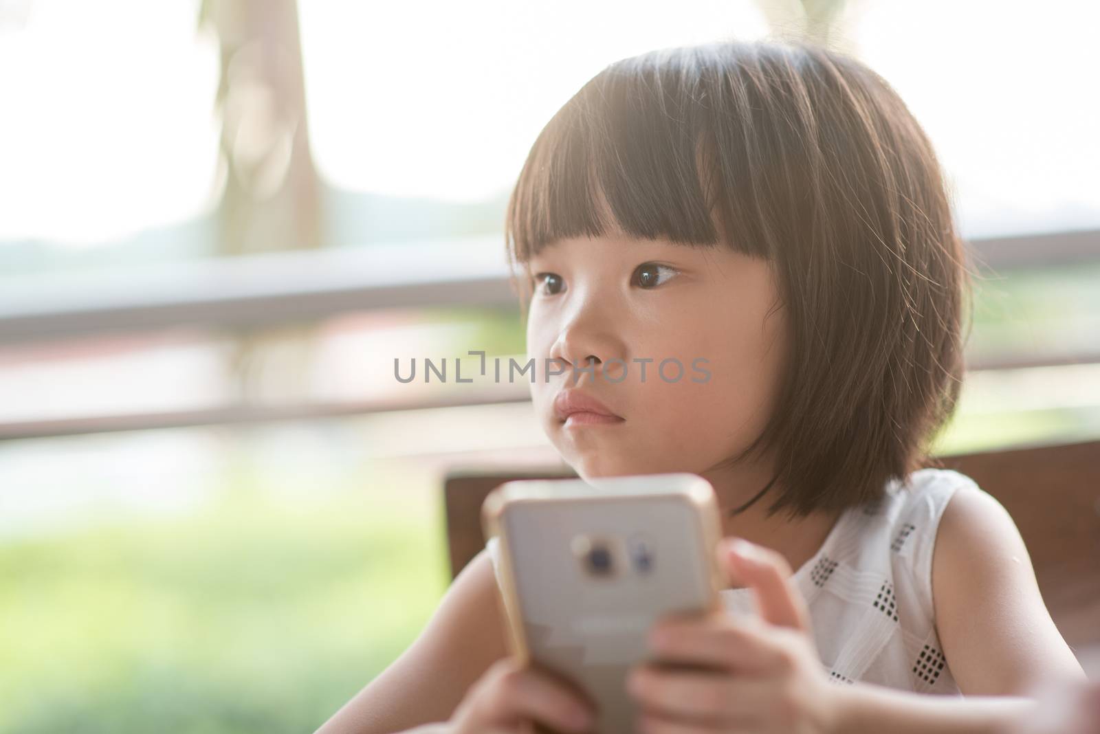 Asian child using smart phone at cafe. Natural light outdoor lifestyle.