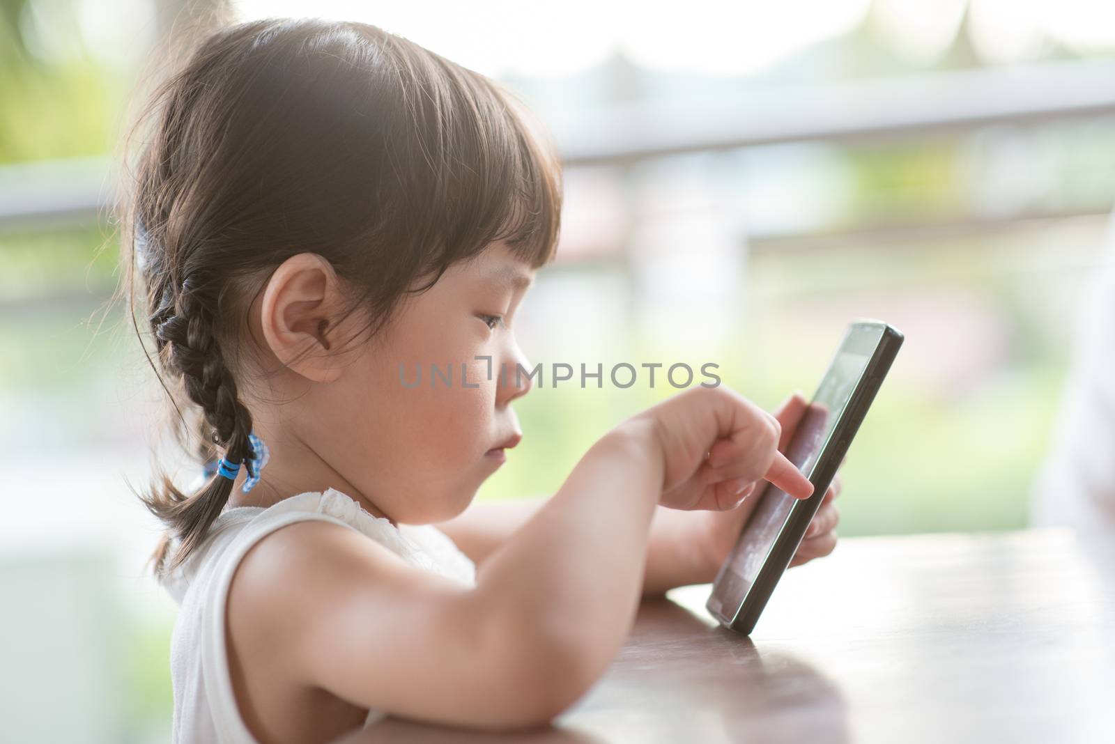 Little Asian girl playing smart phone at cafe. Natural light outdoor lifestyle.