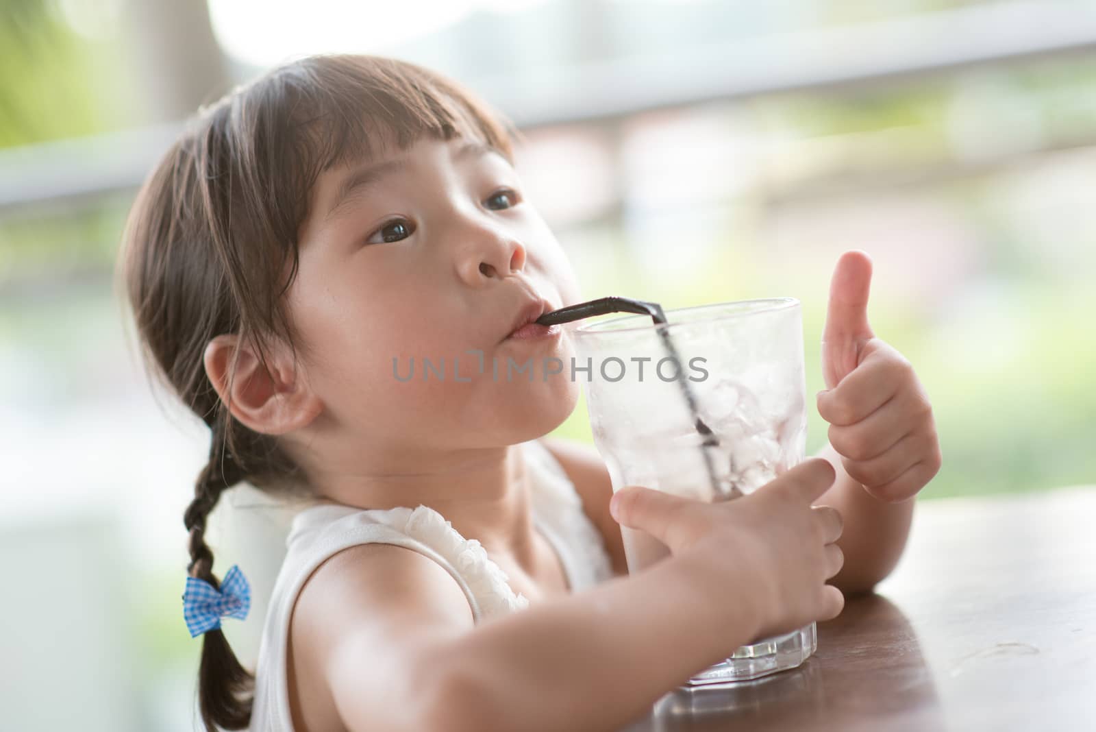 Little Asian child drinking ice chocolate and giving thumb up at cafe. Natural light outdoor lifestyle.