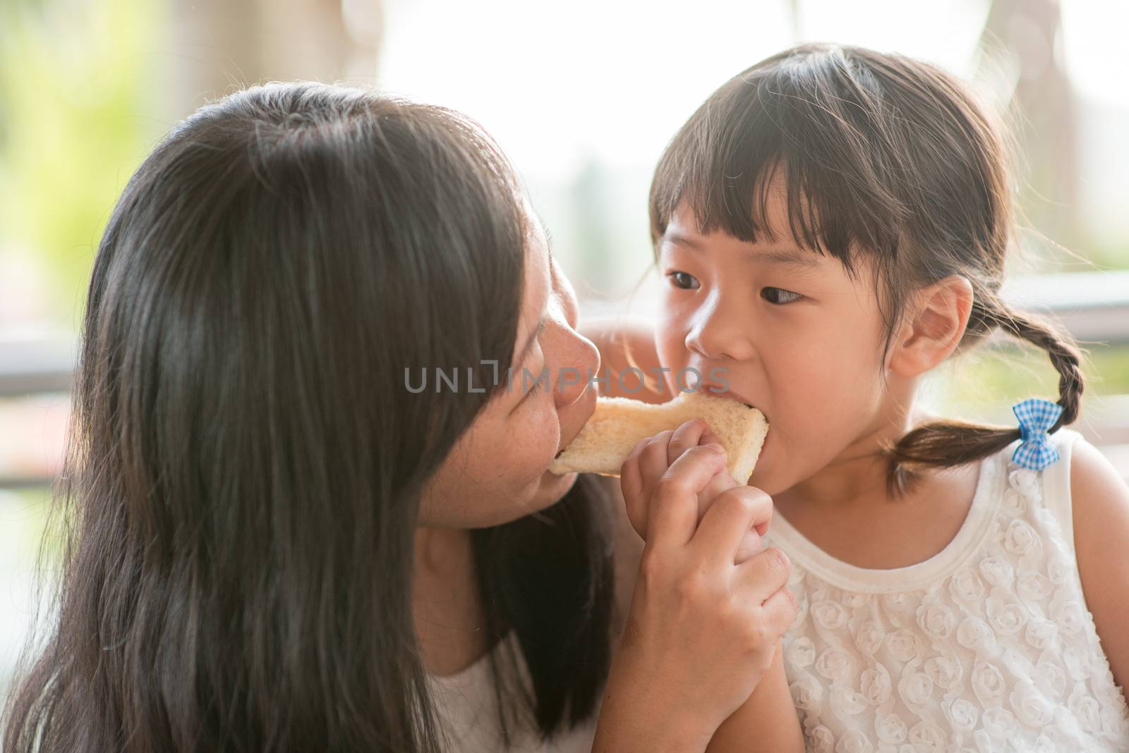 Adorable Asian child eating and sharing butter toast with mom at cafe. Outdoor family lifestyle with natural light.