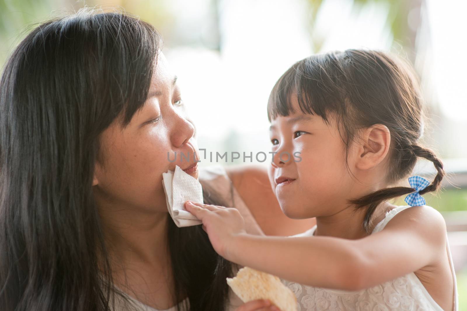 Child wipes mouth for her mom at cafe. Asian family outdoor lifestyle with natural light.