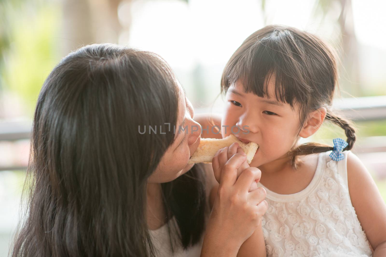 Cute Asian child eating and sharing butter toast with mom at cafe. Outdoor family lifestyle with natural light.