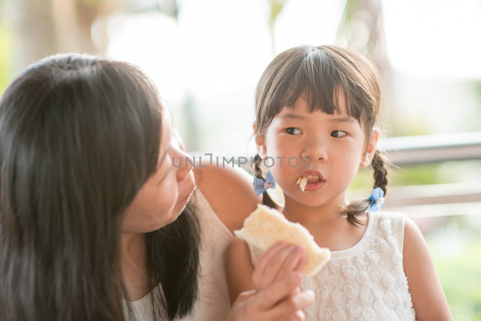 Asian child eating and sharing butter toast with mom at cafe. Outdoor family lifestyle with natural light.