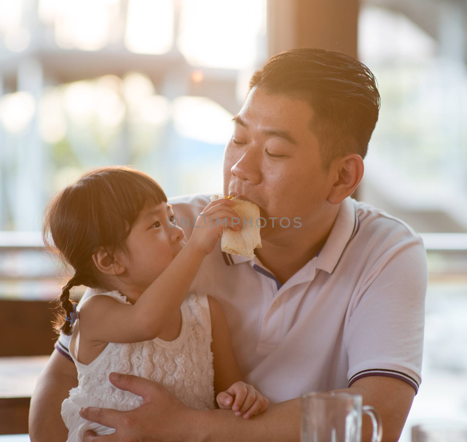 Child feeding bread to daddy at cafeteria. Asian family outdoor lifestyle with natural light.