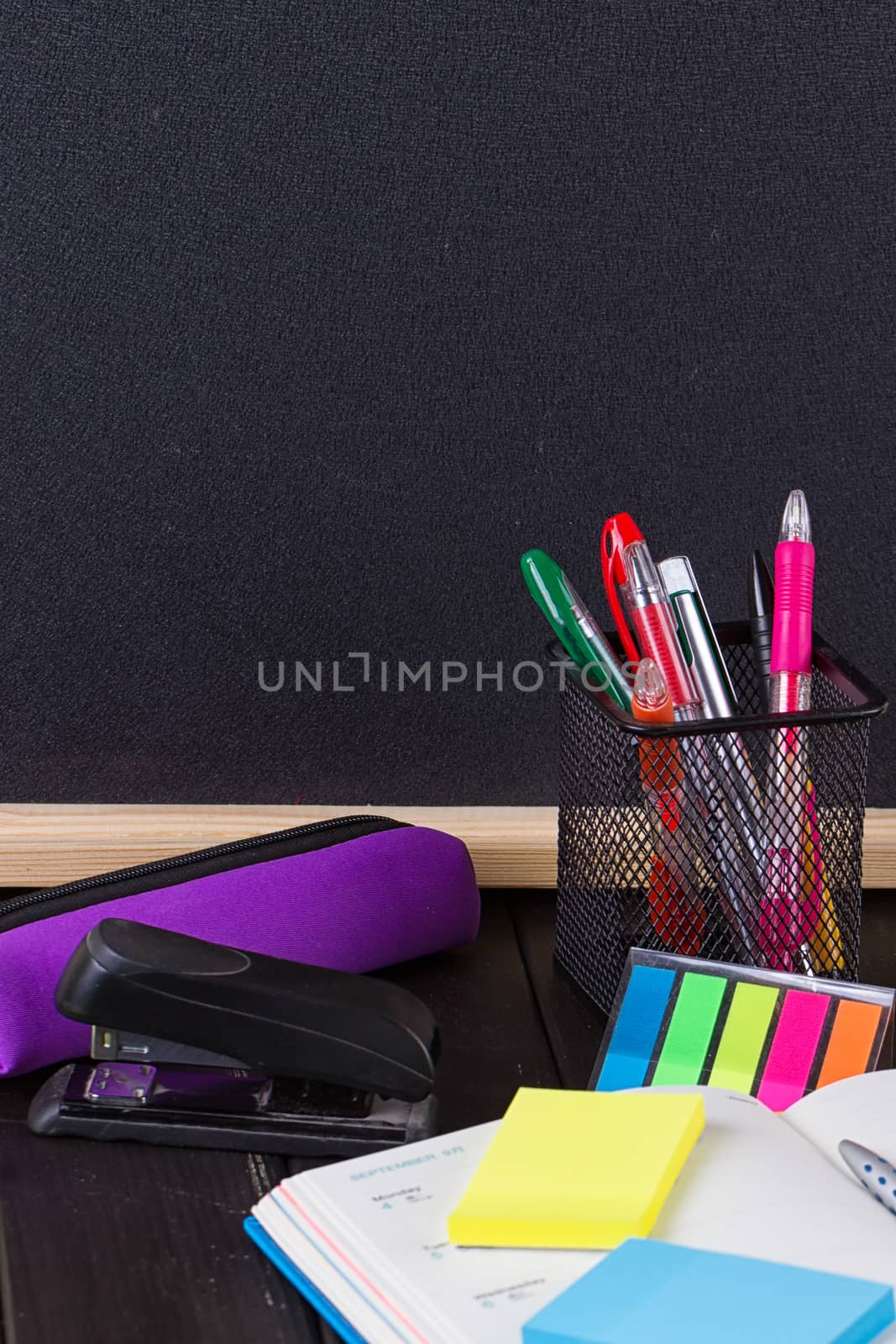 Pencil case with various stationery on old wooden table, on blackboard background