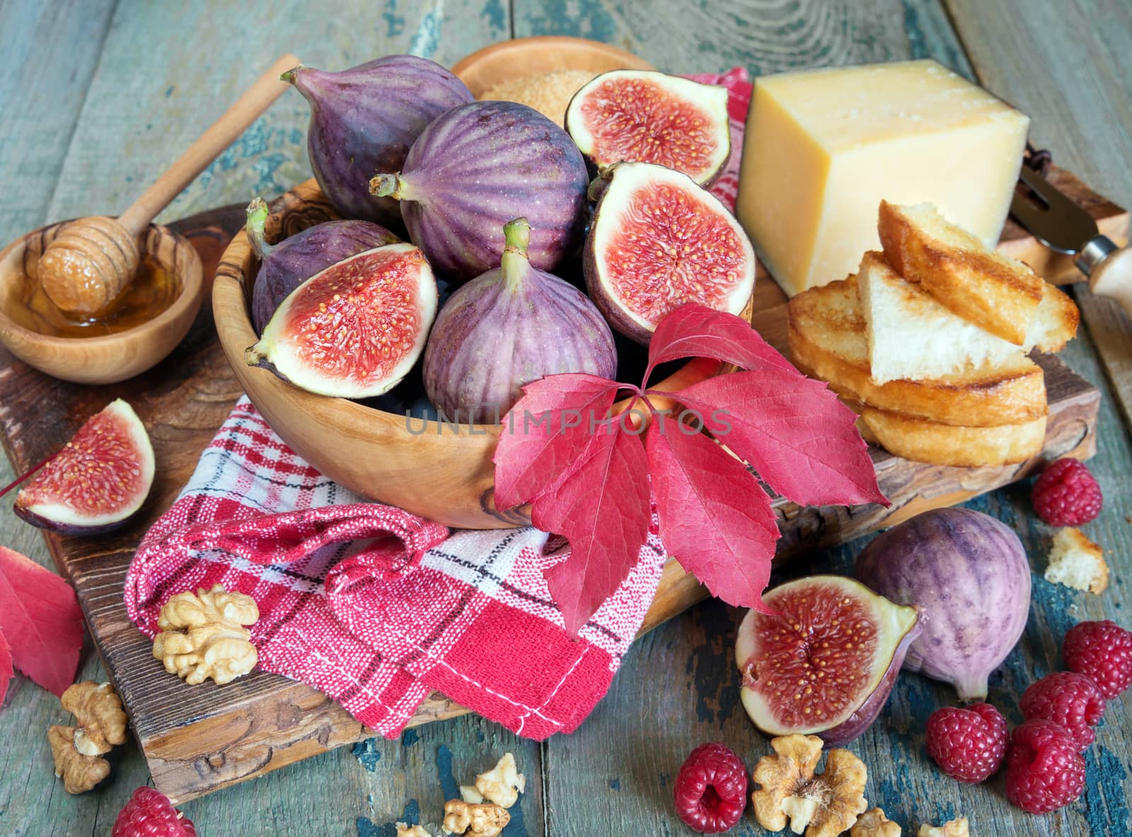 Ripe figs in a wooden bowl, red raspberry, cane sugar, honey and a checkered napkin on old cutting board as well as autumn leaves lie on the old wooden table