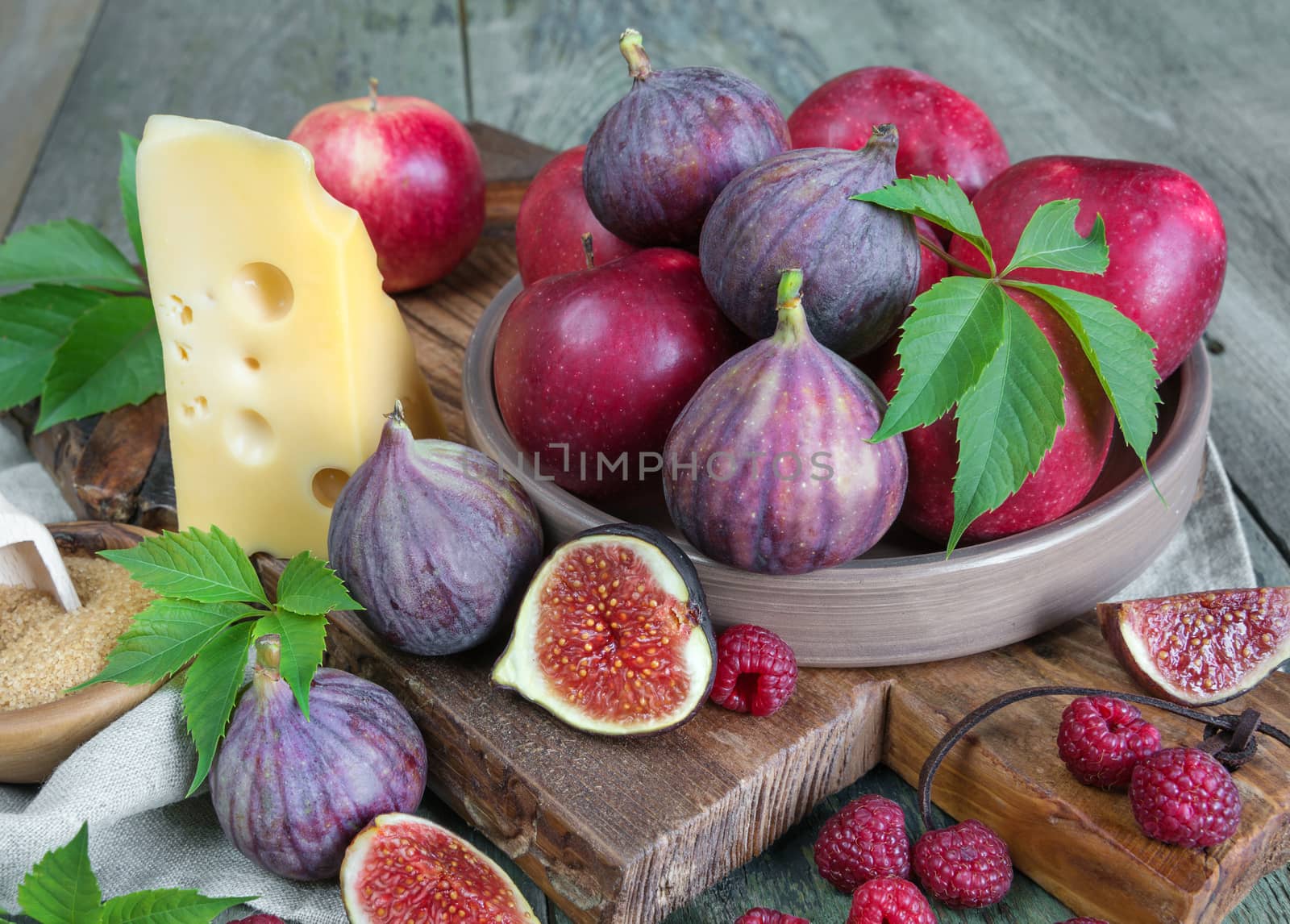Ripe figs, red raspberry and apples, cane sugar and cheese are on old cutting board as well as green leaves lie on the old wooden table