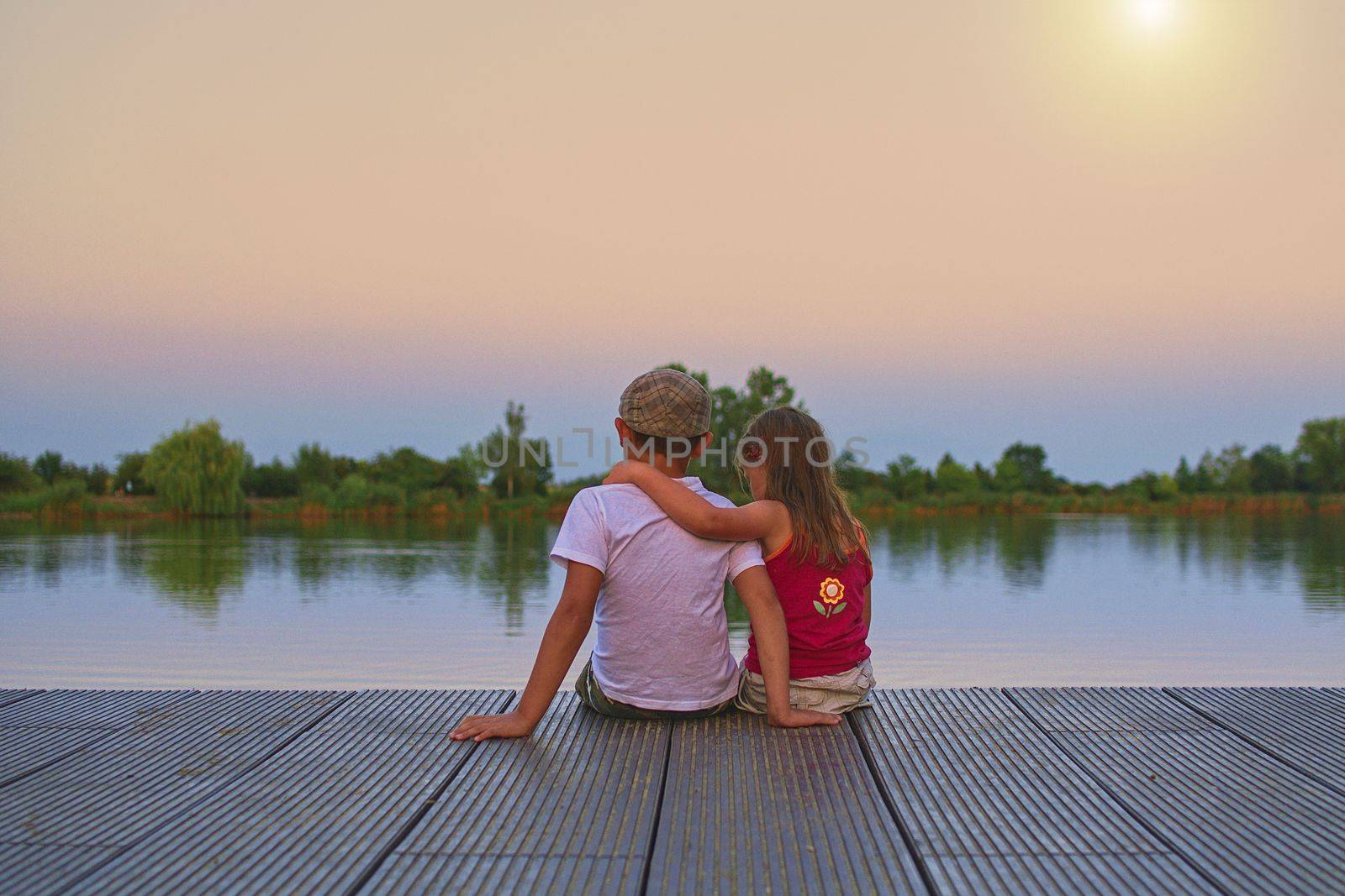 Boy with flat cap and little girl are sitting on pier. Little girl is  a hug her older brother. Love, friendship and childhood concept. Beautiful romantic sunset picture. Copy space.