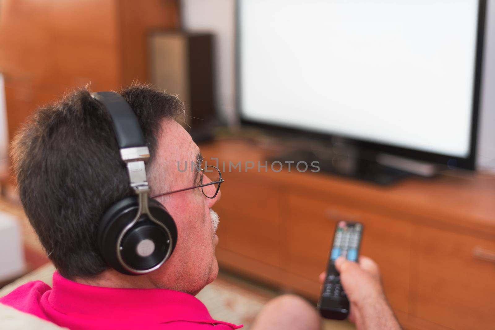 Back view of senior man with headphones watching tv holding remote control