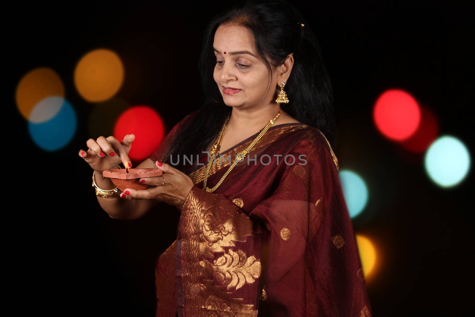 A beautiful Indian woman busy adjusting the flame of the traditional lamp during Diwali festival in India, on the backdrop of blur colorful decorative lights.