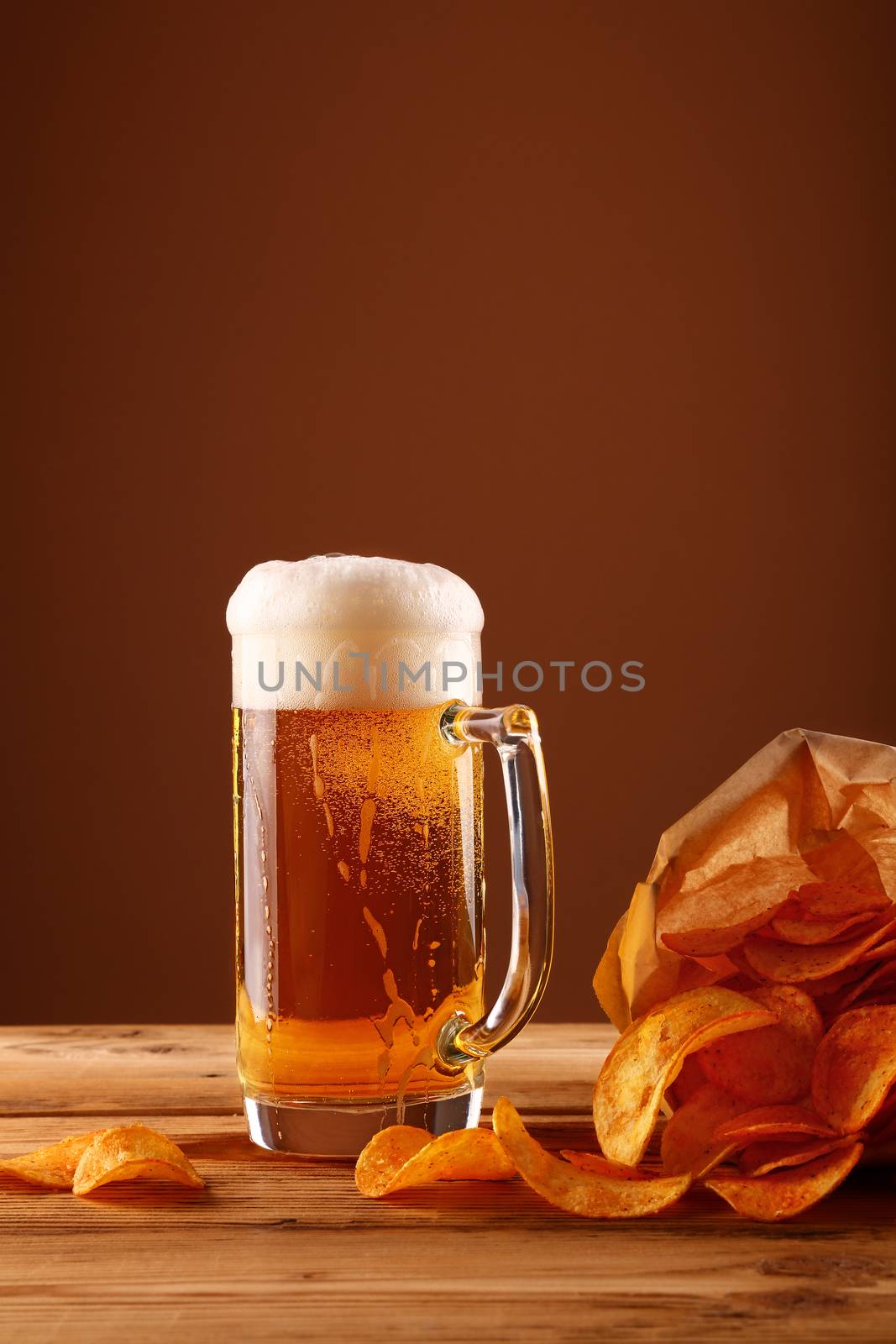 Close up one glass mug of lager beer with white froth and bubbles and paper bag of potato chips on wooden table over dark brown background with copy space, low angle side view