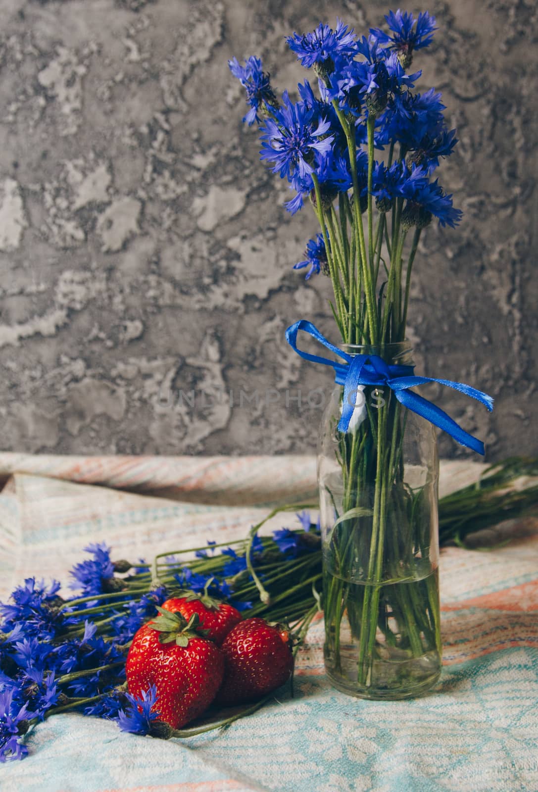 Seasonal summer flowers in vase blue cornflowers and fruits strawberries on a napkin close-up conceptual background