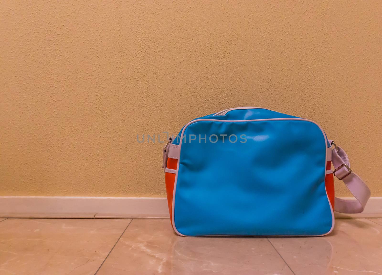 blue with red messenger school bag isolated in against a wall and standing on the floor in a room by charlottebleijenberg