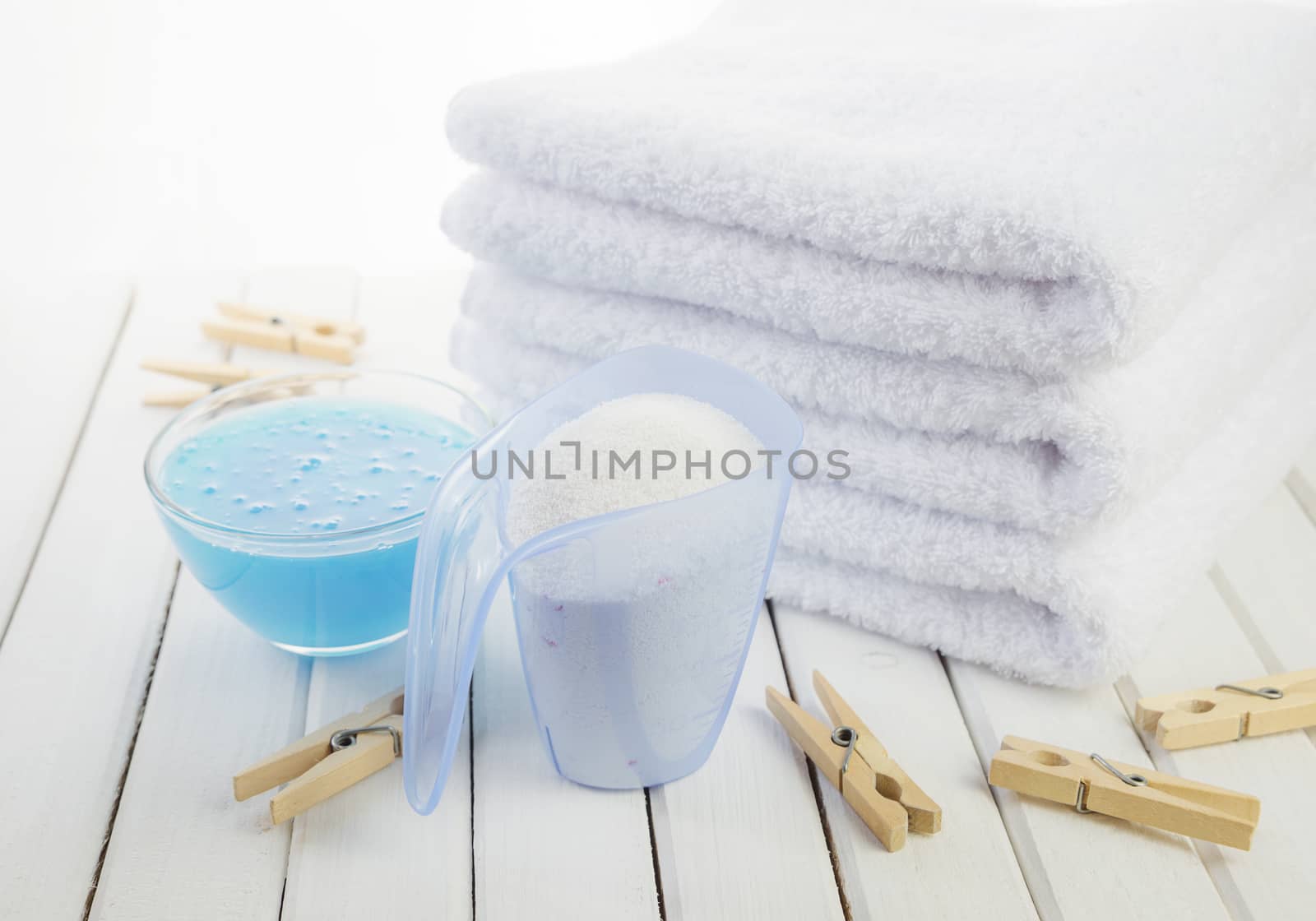 Stack of three white fluffy bath towels, washing powder in measuring cup, blue fabric softener in a transparent glass bowl and wooden clothespins on the background of white boards