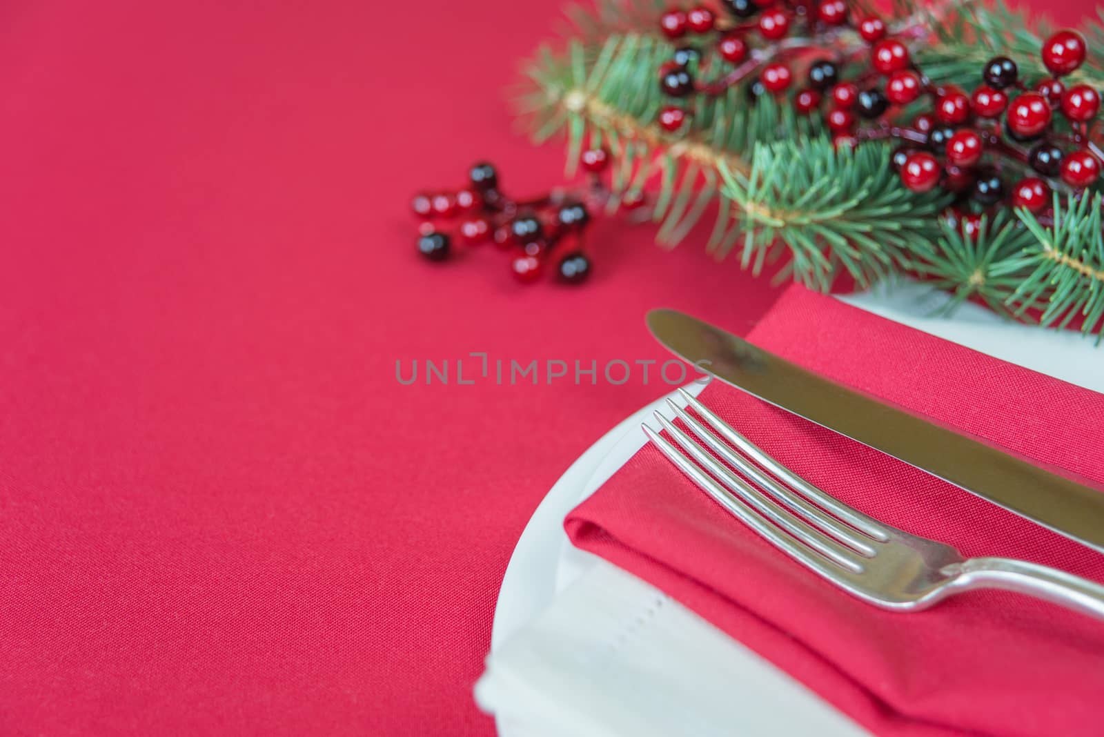 Silver knife and fork lie on the red linen napkin, as well as red holly berries and green spruce branch, which is located on a table covered with a red tablecloth, with space for text