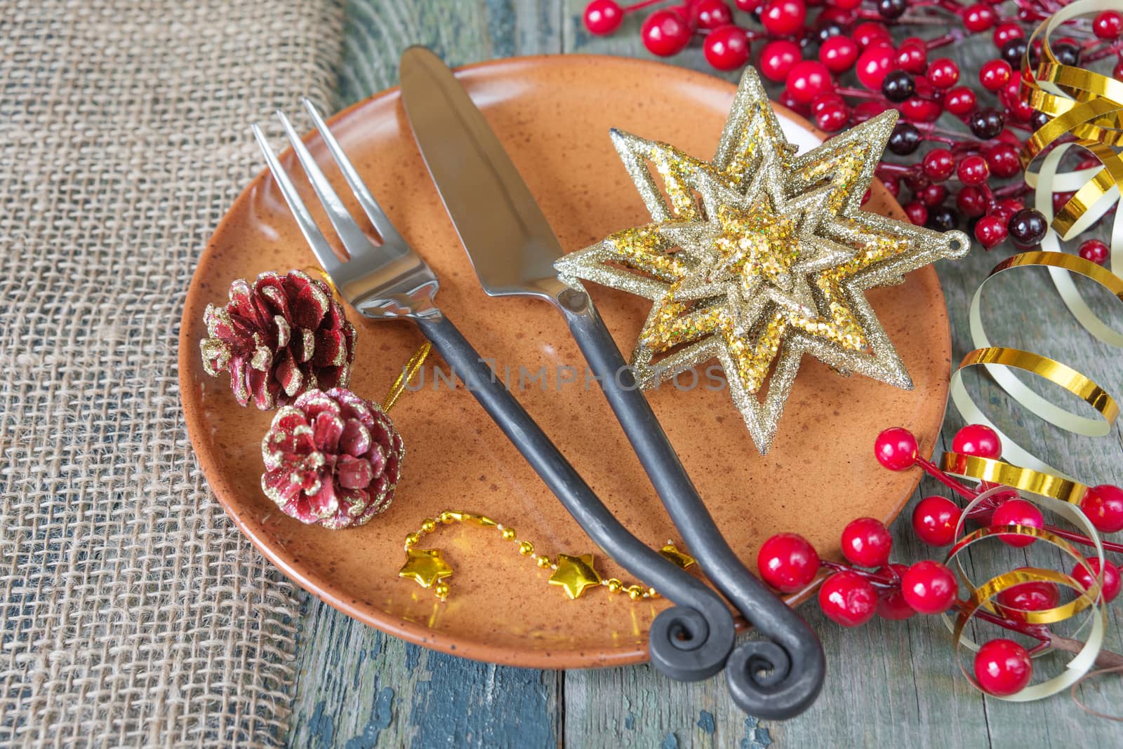 Beautiful Christmas table: handmade still  knife and fork are on the brown ceramic plate, golden star and ribbon, red holly berries which is located on a old wooden boards