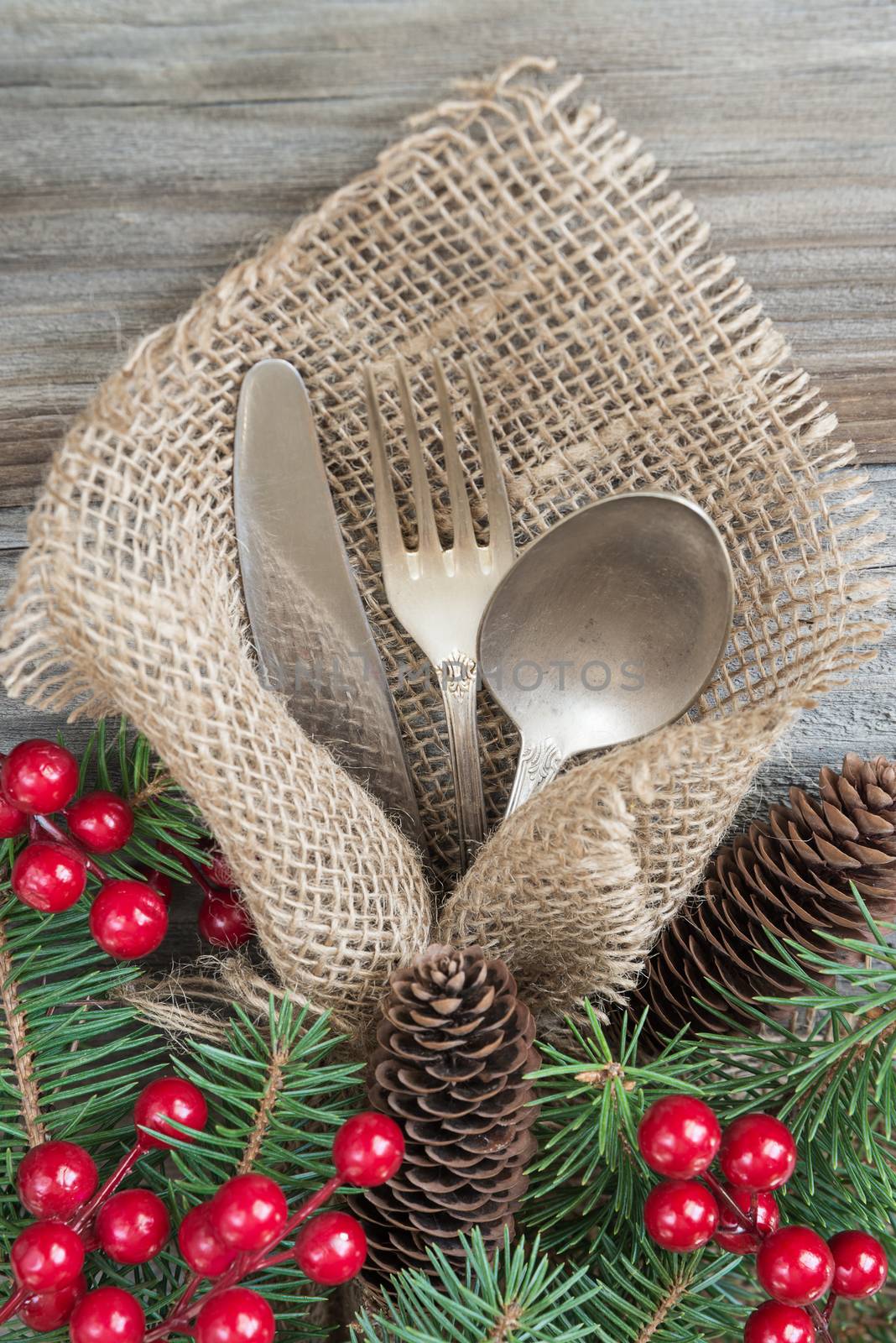 Christmas table with Christmas decoration: old knife, spoon and fork lie on the sacking napkin, as well as red holly berries and green spruce branch, which is located on an old wooden table