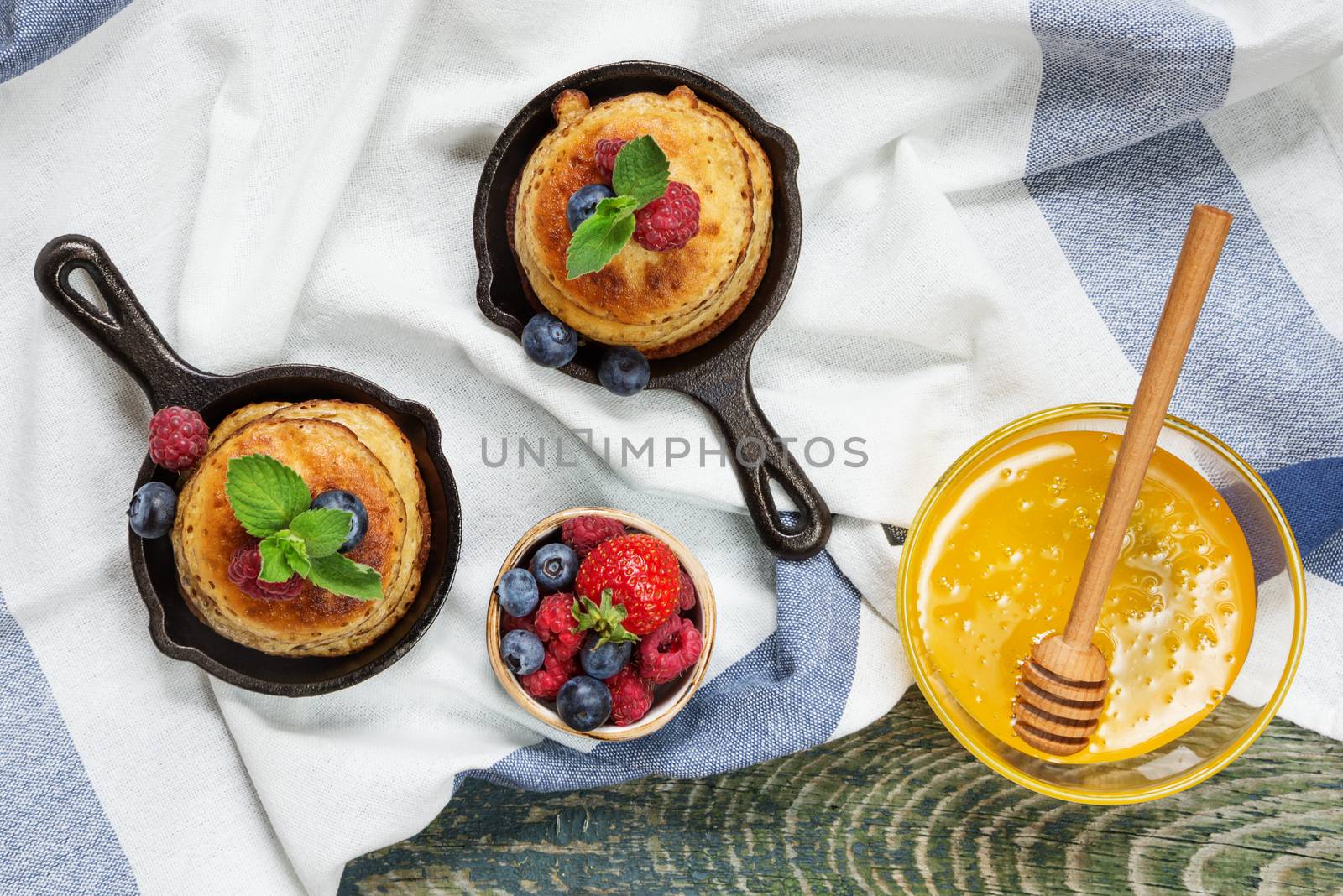 Pancakes in cast-iron frying pans with fresh berries and honey in a glass bowl on the background of the old wooden boards covered with a linen tablecloth