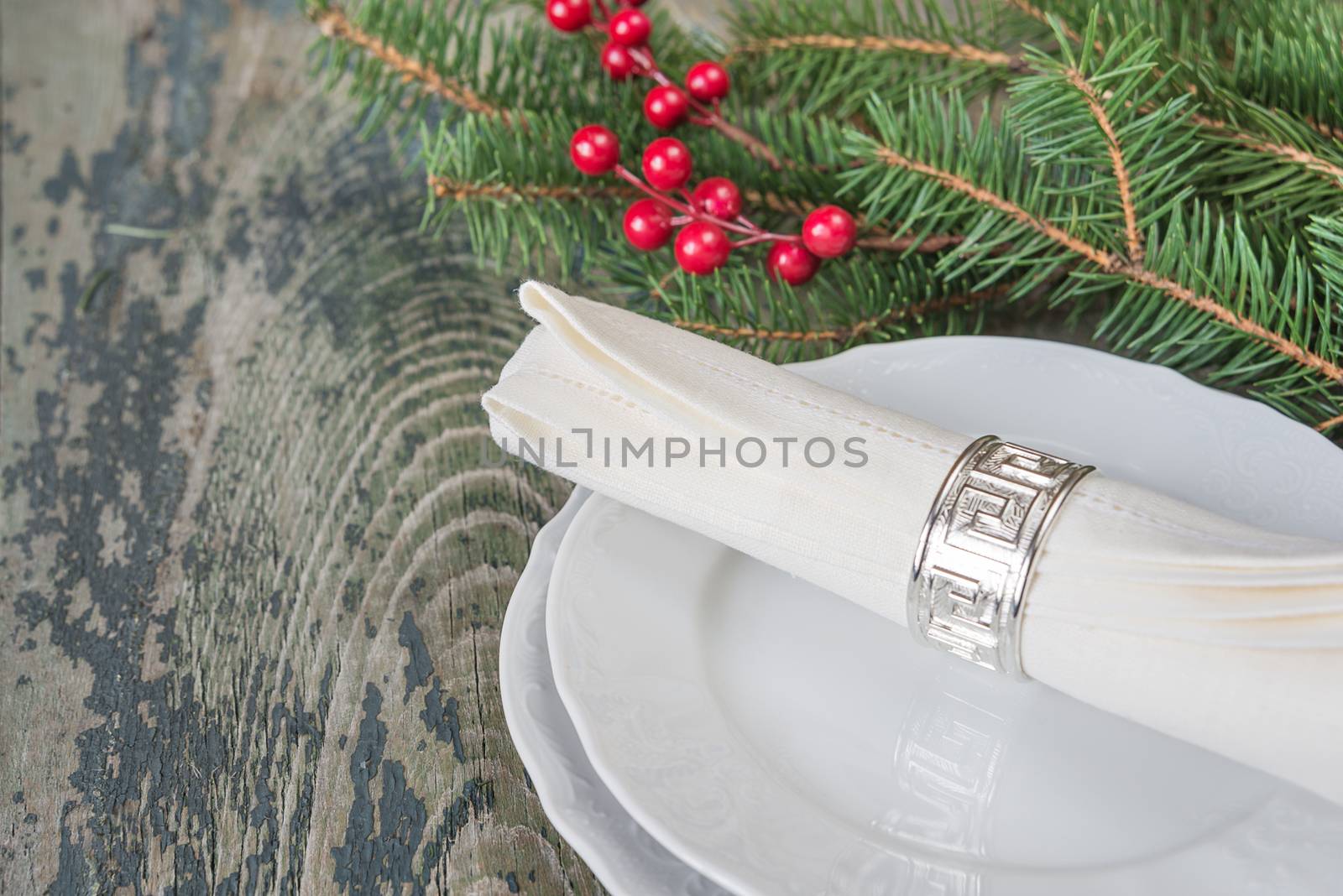 White linen napkin, silver napkin ring are on the white dinner plate, as well as red holly berries and green spruce branches which is located on a old wooden table, with space for text