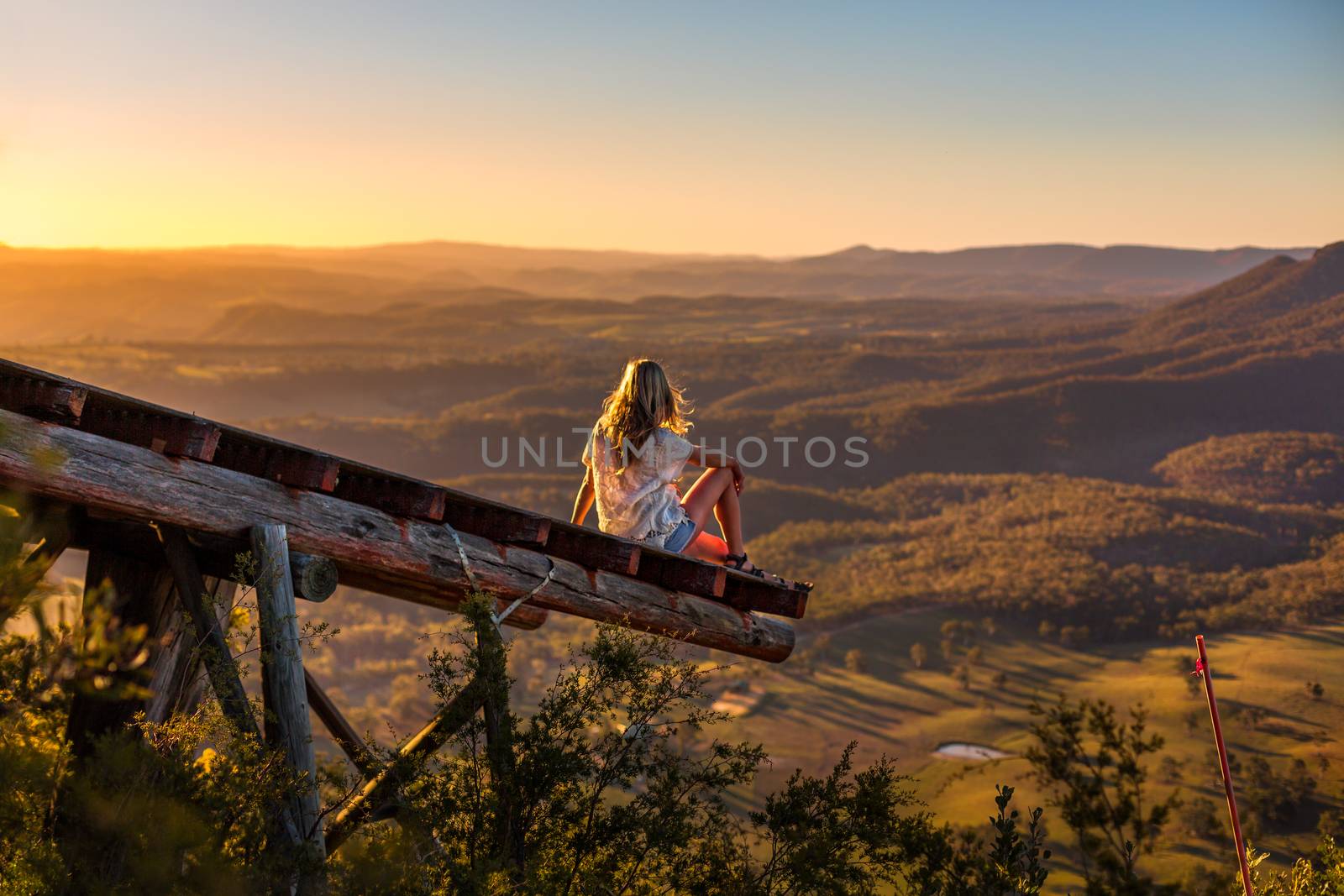 Female in casual clothes with sunlit hair flowing in the breeze relaxes on the timber platform that extends out over the valley and watches the golden sunlight fill the valley as the sun sets in the west.