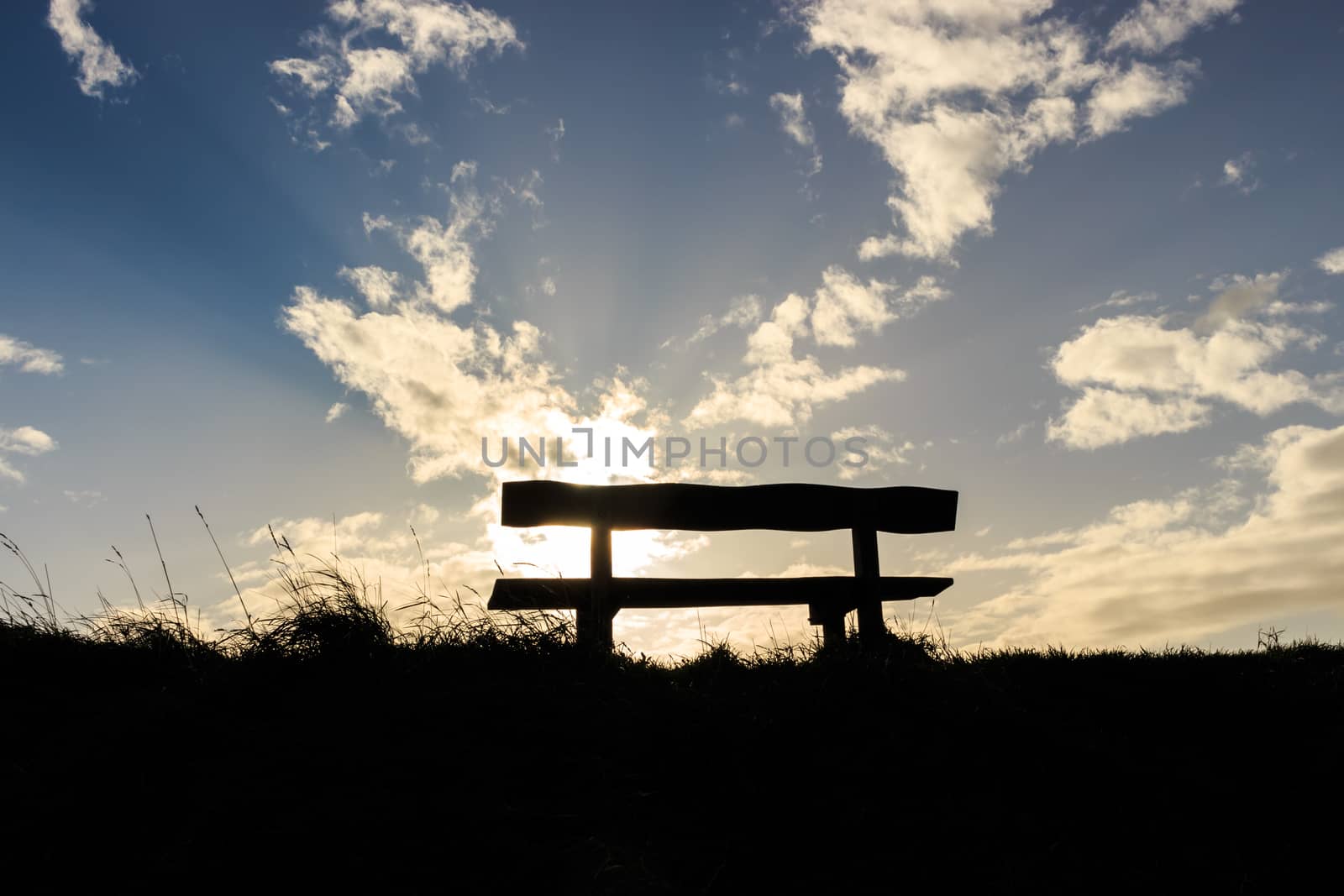 Thoughtful and peaceful silhouette wooden bench and grass with a blue and white soft fluffy sky in the background, light rays