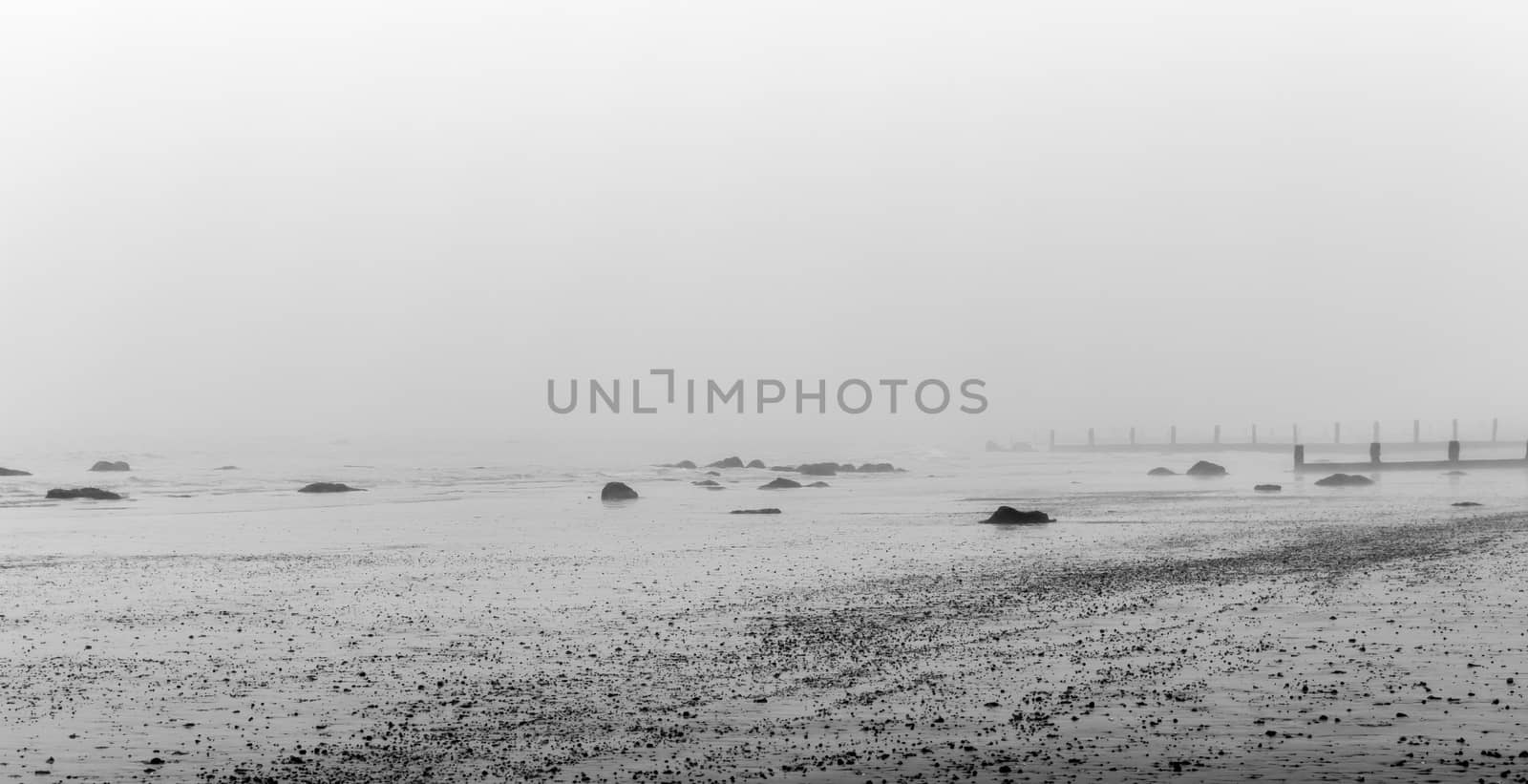 Monochrome Misty beach in the winter with rocks in the foreground and sea breaks behind