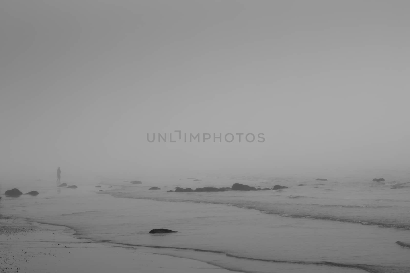 single person on a beach with waves on sand and rocks in the background in monochrome