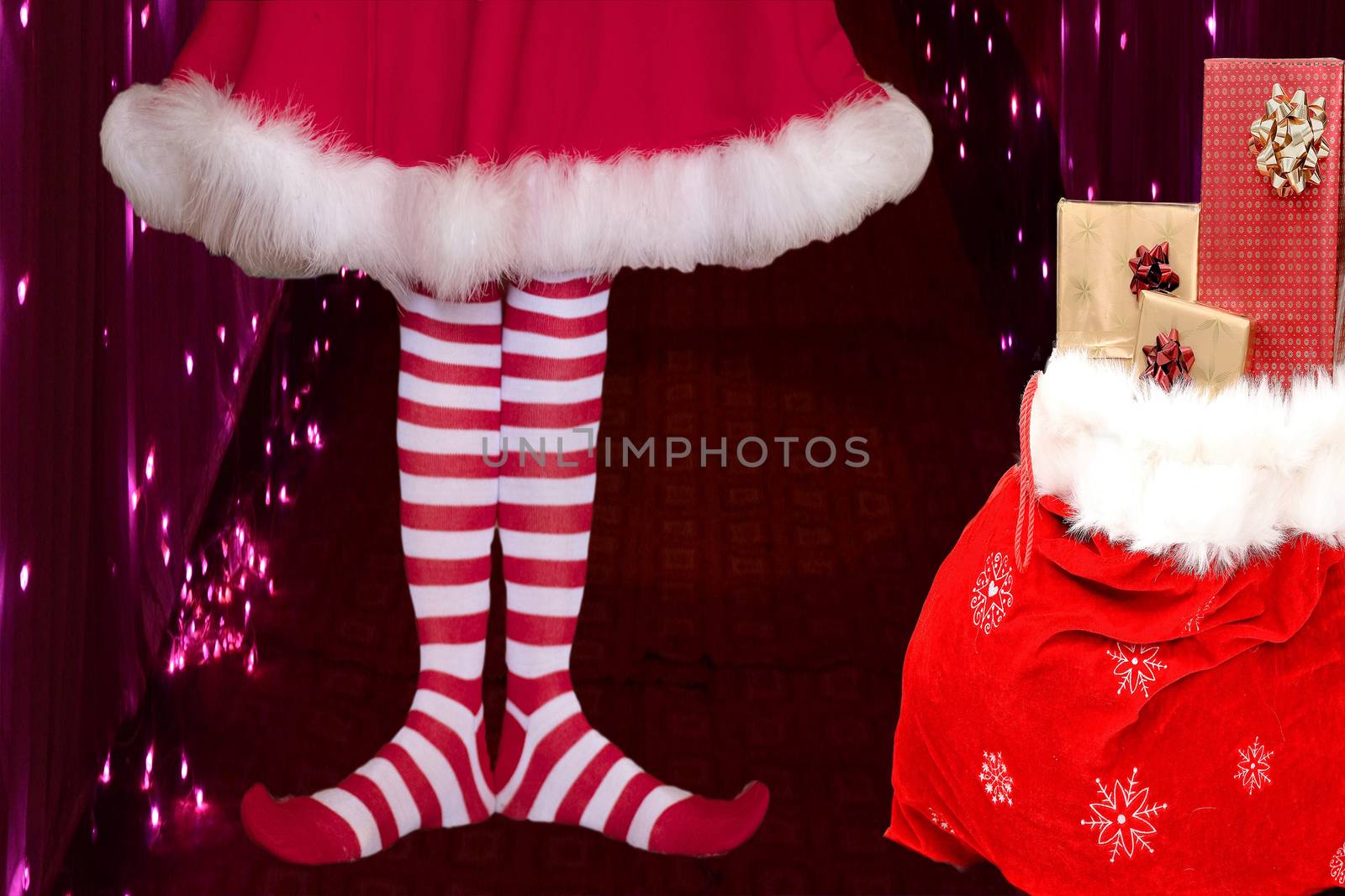 cute girl wearing red white striped elf stockings and a cute dress standing next to a santa claus bag full of presents on a background with christmas lights
