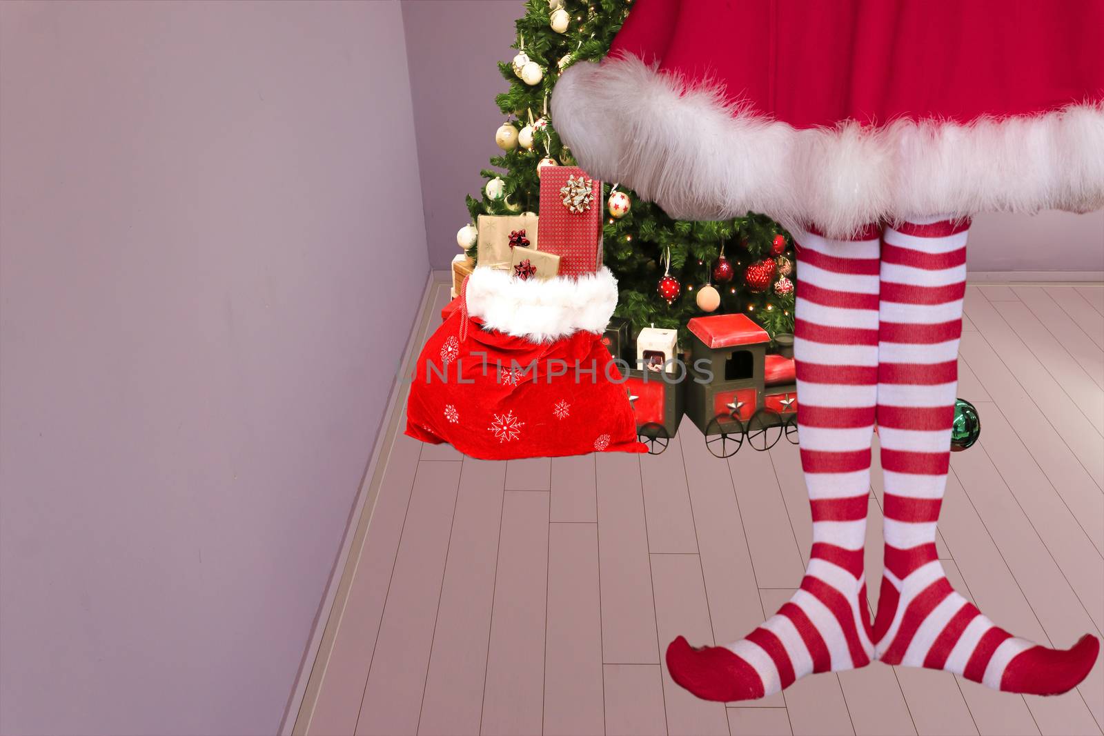 cute girl with pointed elven feet wearing elf legging standing in a room with a christmas tree and santa claus bag full of presents on the background