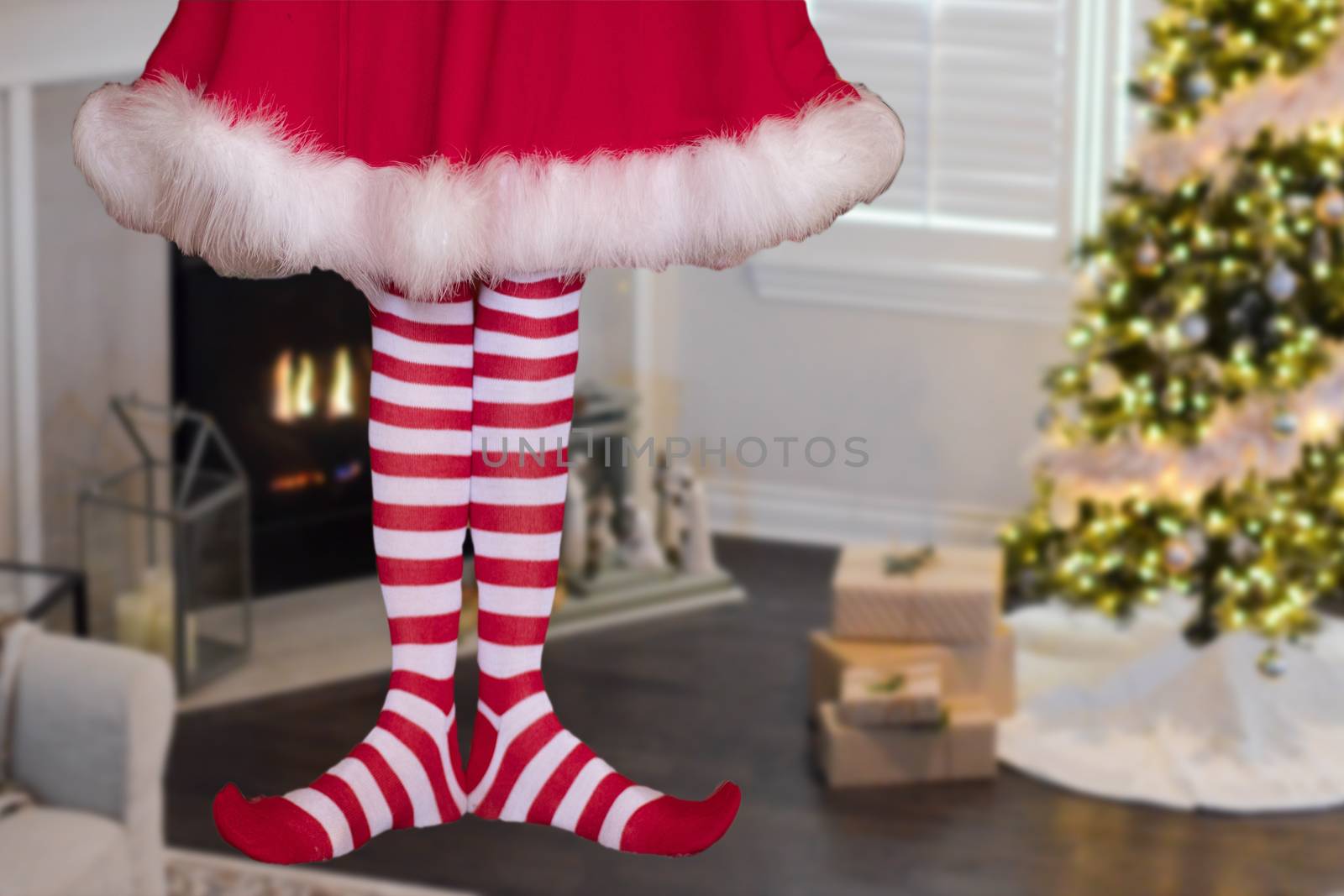 cute little christmas elf girl wearing striped stockings standing in a decorated christmas living room with gifts and christmas tree in the background